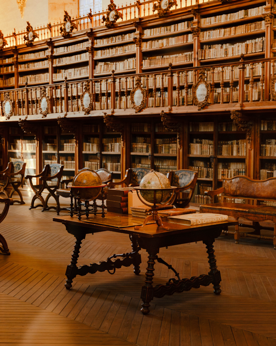 Une bibliothèque ancienne avec des étagères en bois remplies de livres, des globes terrestres sur une table en bois, et des fauteuils en bois et tissu.