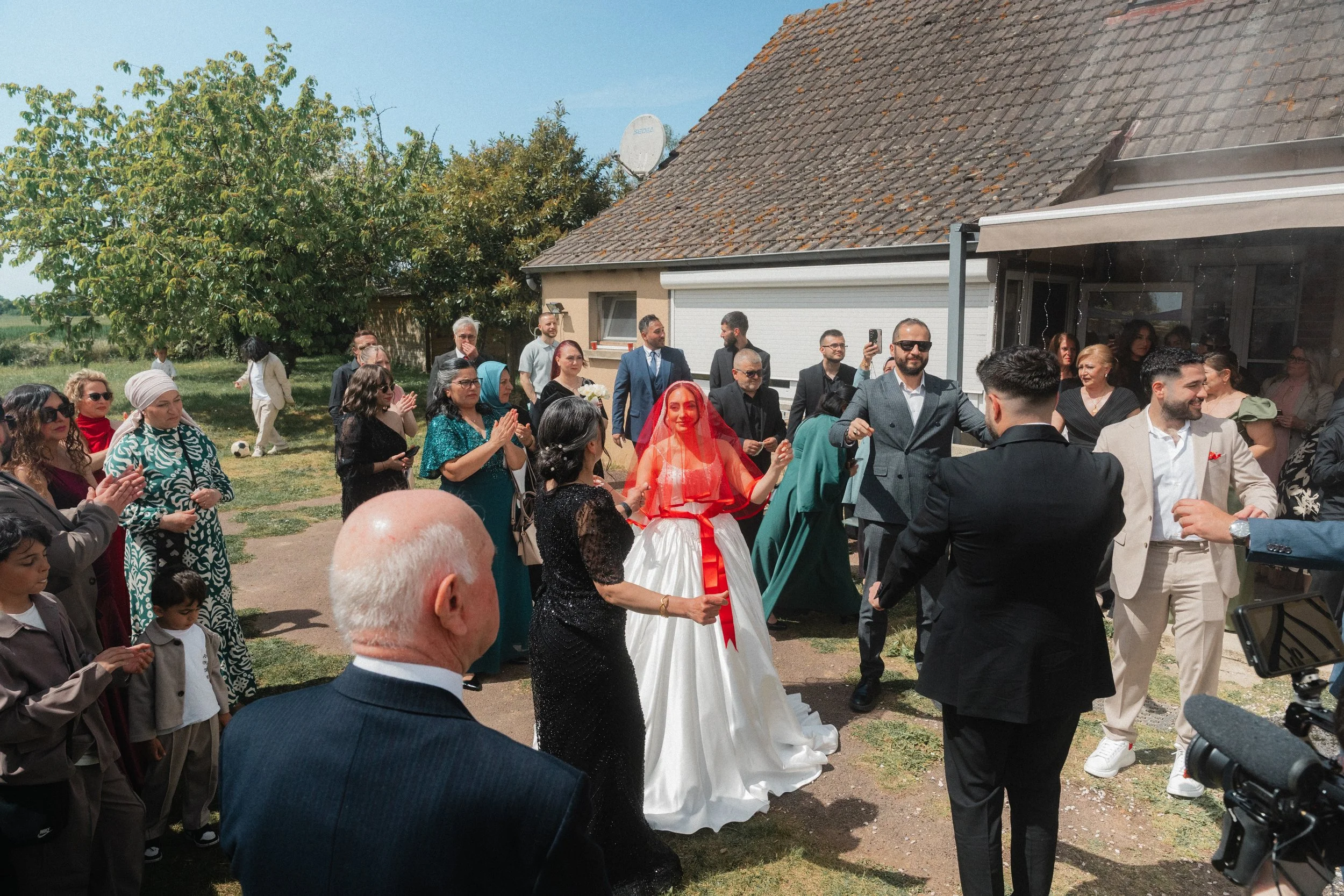 Groupe de personnes lors d'une célébration en plein air, certaines portant des tenues de cérémonie comme une mariée en robe blanche avec un voile rouge, d'autres en costumes, dans un jardin avec une maison en arrière-plan.