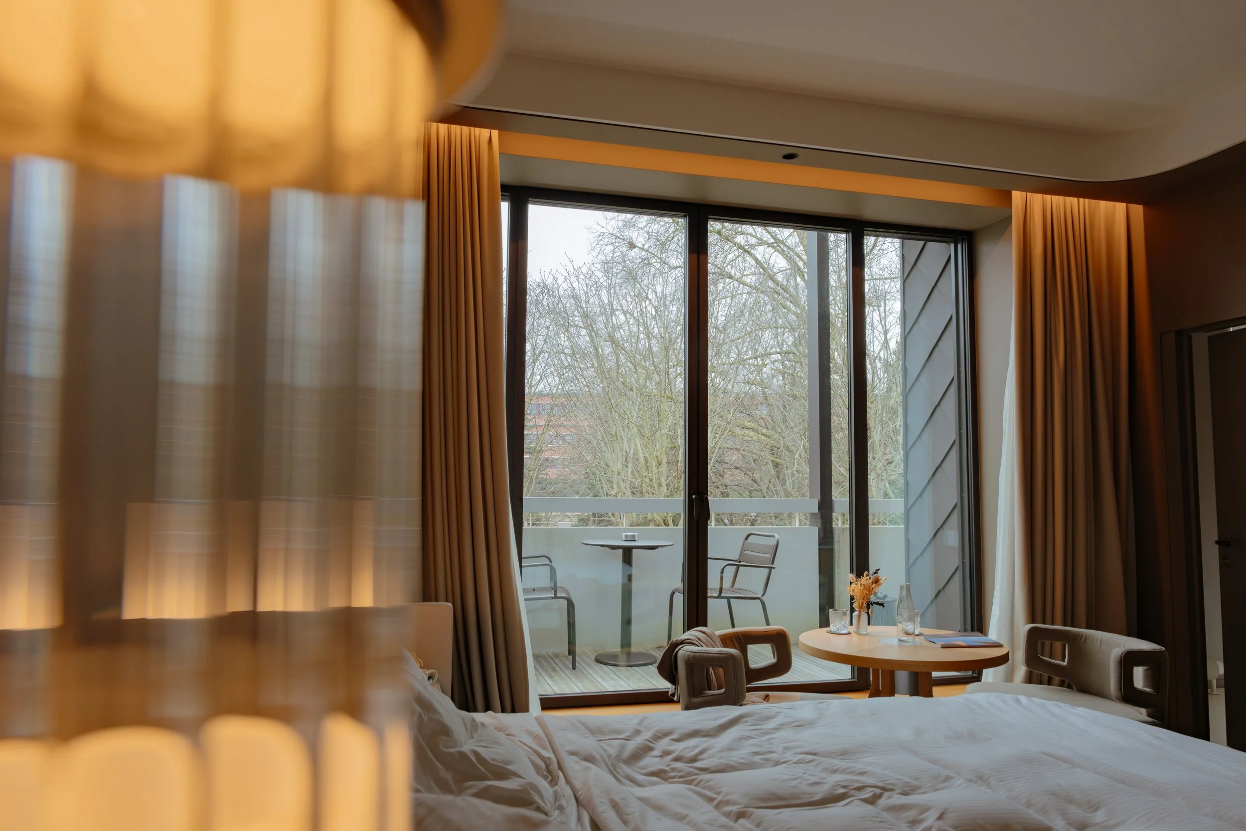 Chambre avec grand mur de fenêtres en verre, vue sur un balcon avec une table et deux chaises, rideaux dorés, décor moderne