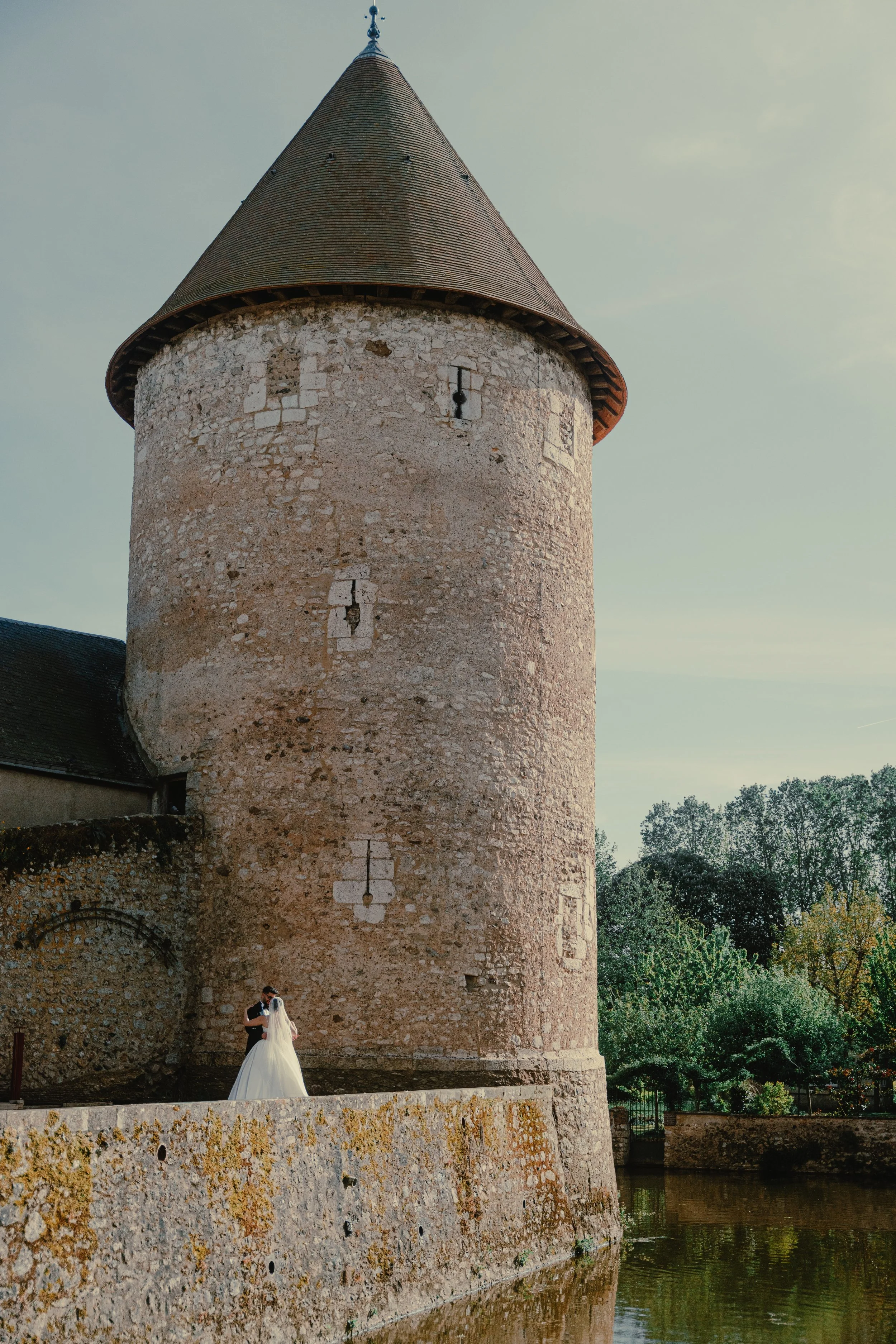 Un couple de mariés se tenant par la main devant un vieux château en pierre avec une tour conique en arrière-plan, près d'un plan d'eau, le tout sous un ciel clair.