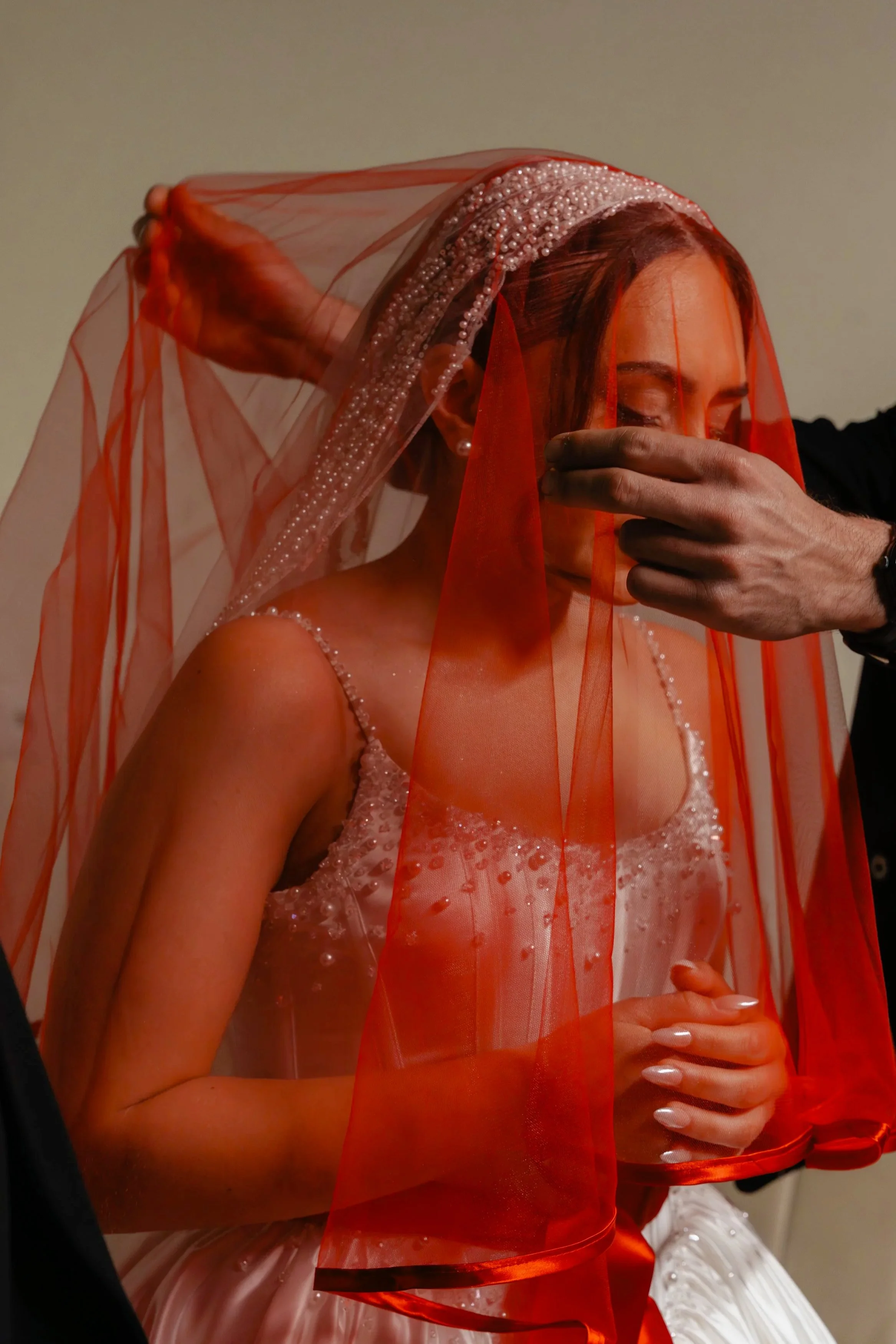 Une femme en robe blanche avec de nombreux perles et un voile rouge tient ses mains jointes devant son abdomen, une autre personne ajuste le voile sur sa tête.