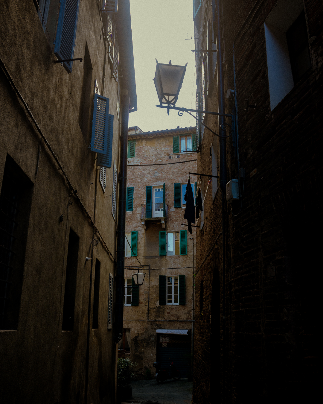 Vue d'une ruelle étroite entre des bâtiments en pierre avec des volets bleus, une lanterne suspendue et des vêtements suspendus pour sécher.