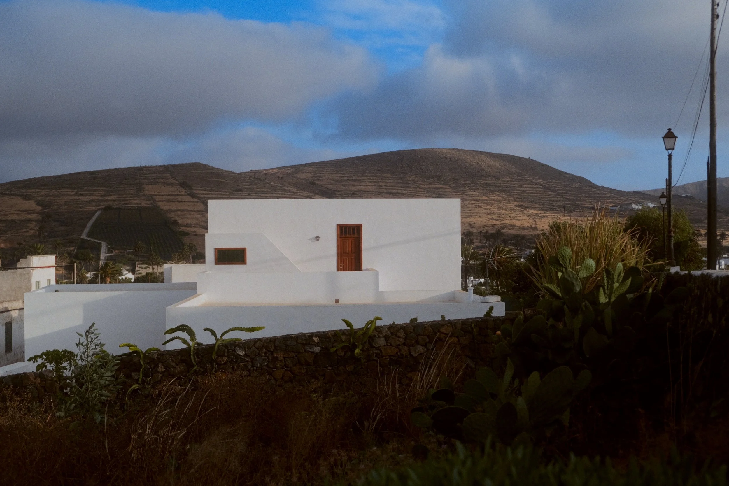 Une maison blanche moderne avec une porte en bois, au sommet d'une colline, avec un ciel nuageux et des lampadaires le long du chemin. Des plantes succulentes et un mur de pierre sont visibles au premier plan.