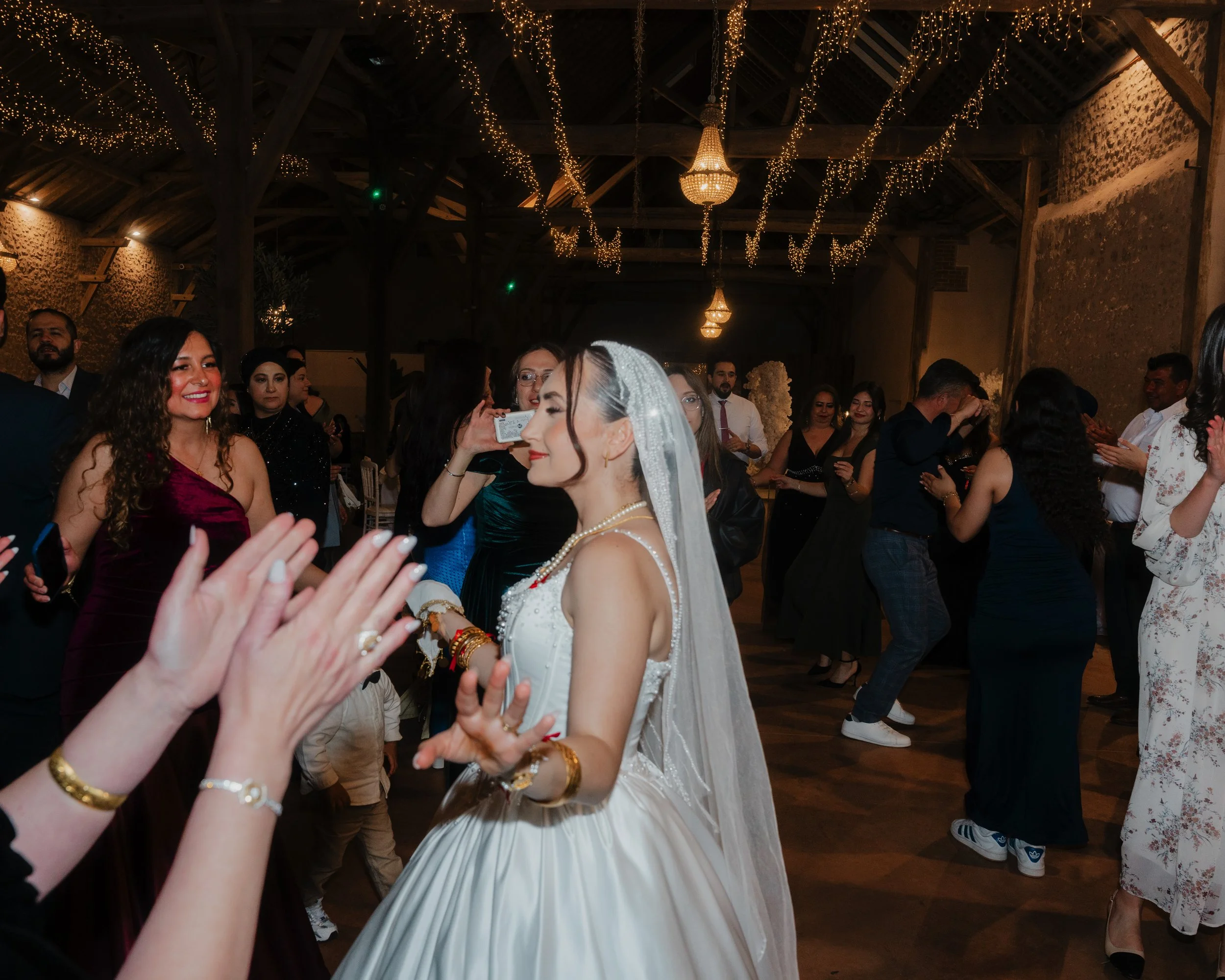 Groupe de personnes lors d'une célébration, probablement un mariage, dans une salle décorée avec des guirlandes lumineuses et un lustre chandelier, où une femme en robe de mariée est visible au centre.