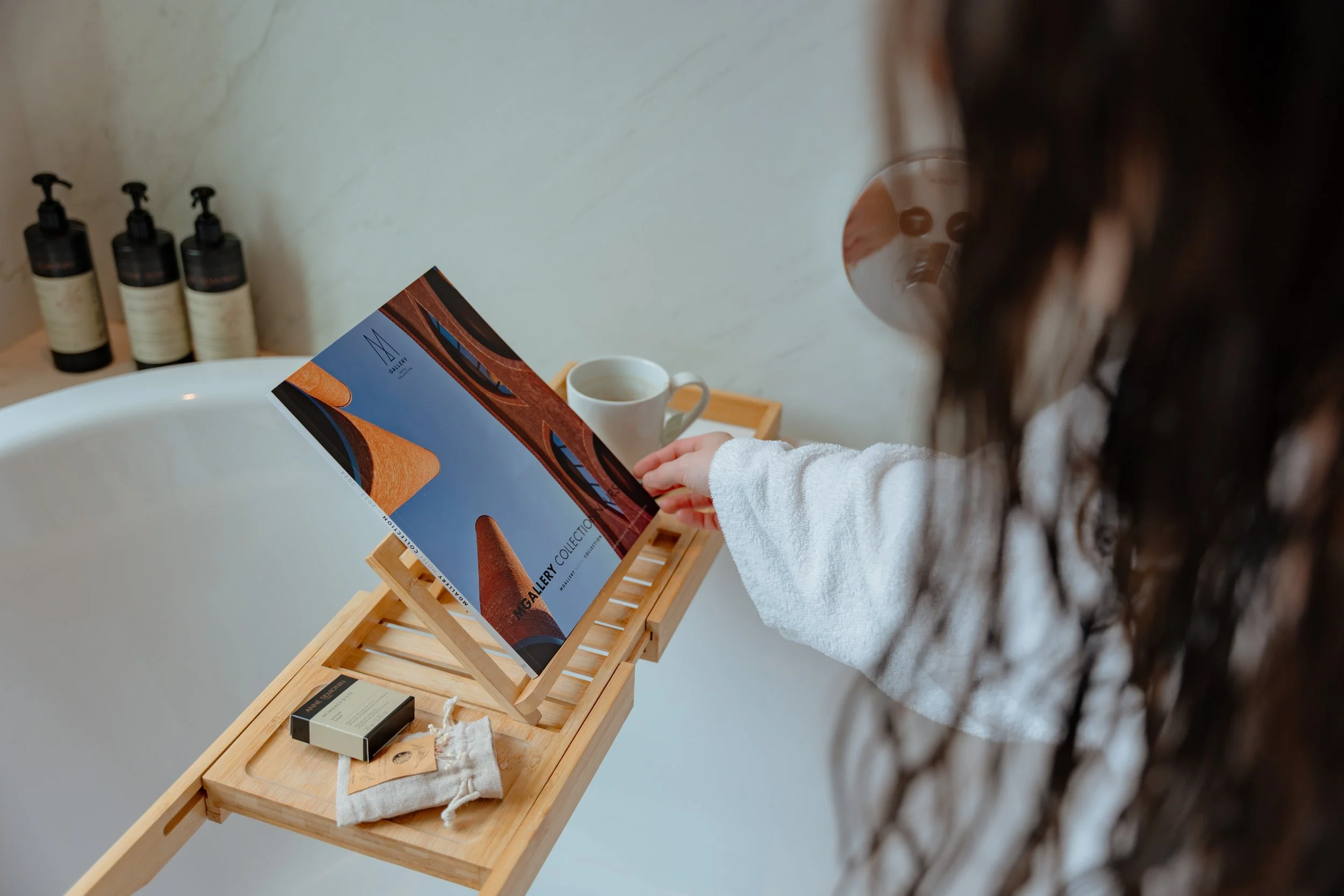Une personne regarde un catalogue d'art dans une boutique ou une galerie, avec des bouteilles de savon ou lotion en arrière-plan et un mug. La personne porte un vêtement blanc à manches longues. La boutique a un meuble en bois clair et un miroir rond en arrière-plan.