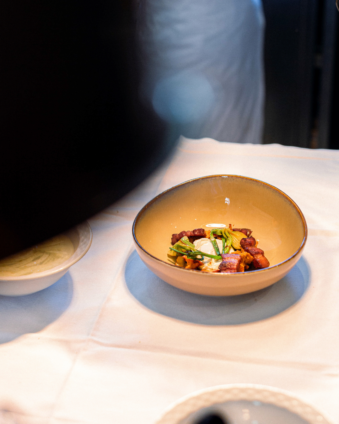 Plat de nourriture dans un bol beige contenant des morceaux de viande, de la crème, et des légumes verts, posé sur une nappe blanche.