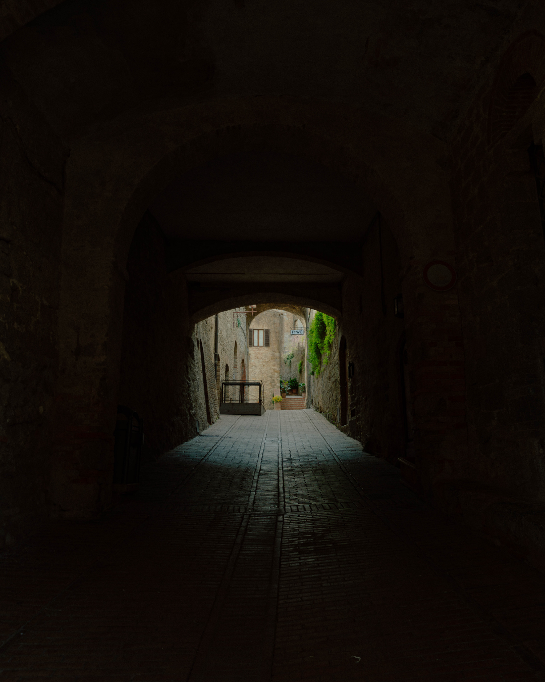 Vue à travers une passage souterrain menant à une petite place pavée avec des plantes en pot, bâtiments en pierre et un panneau indiquant des toilettes.