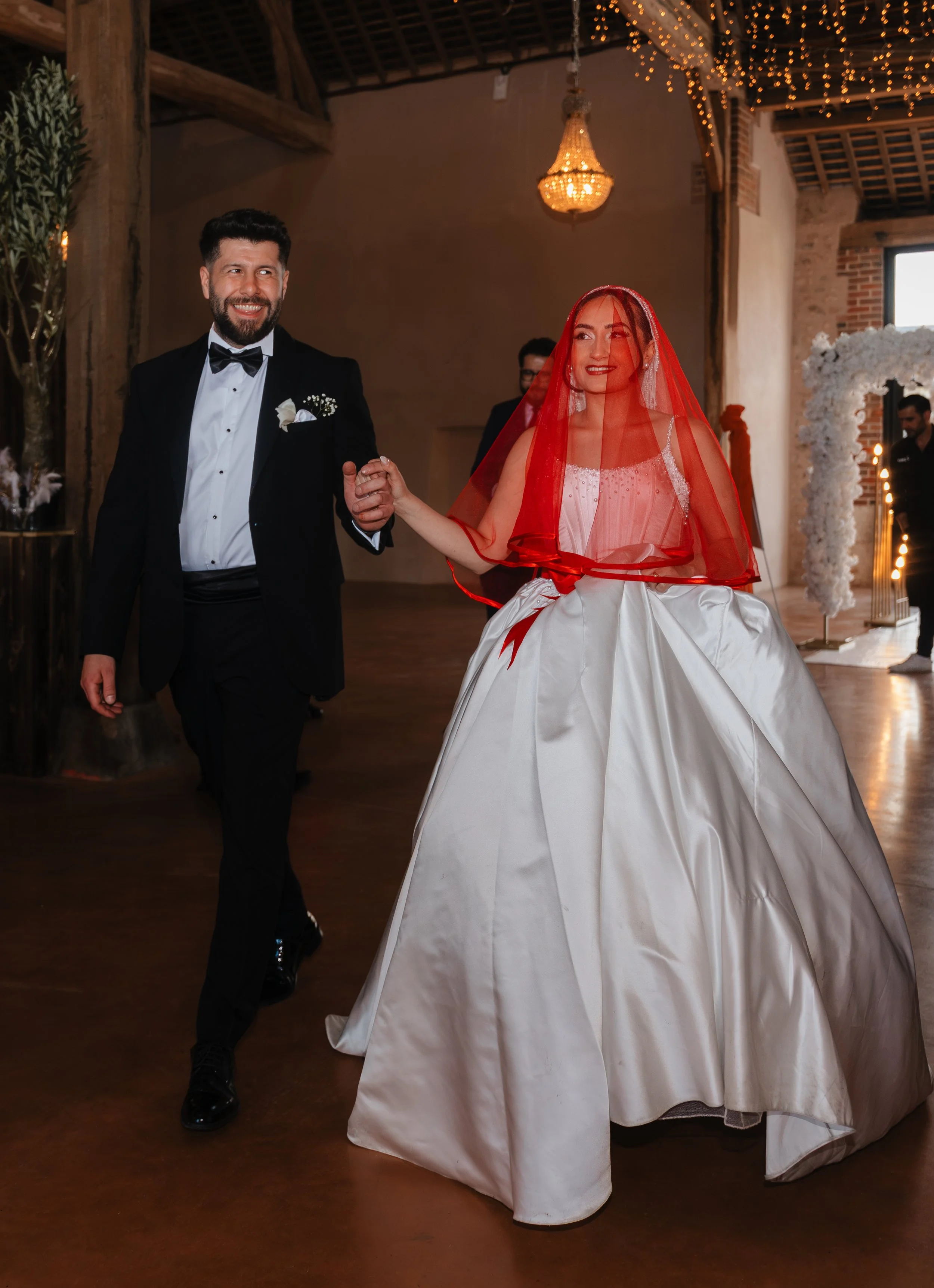 Un couple marié, le homme en costume de smoking noir et la femme en robe de mariée de style vintage avec une voilette rouge, dansant lors d’un mariage dans une salle décorée avec des guirlandes lumineuses et une arche de fleurs en toile de fond.