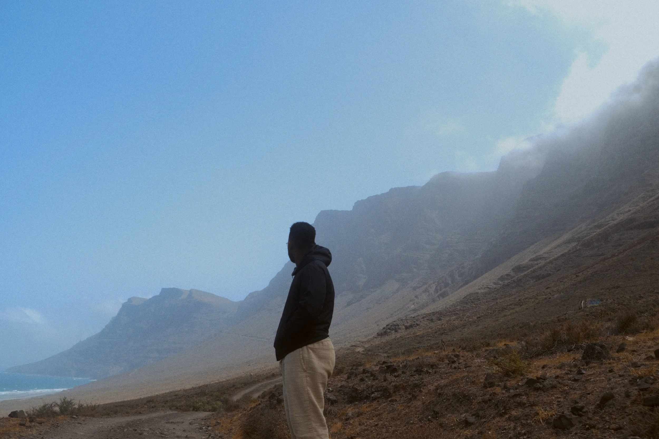 Un homme se tient sur une plage rocheuse avec des montagnes en arrière-plan, vêtu d'un manteau et regardant vers le haut, face à un ciel partiellement nuageux.