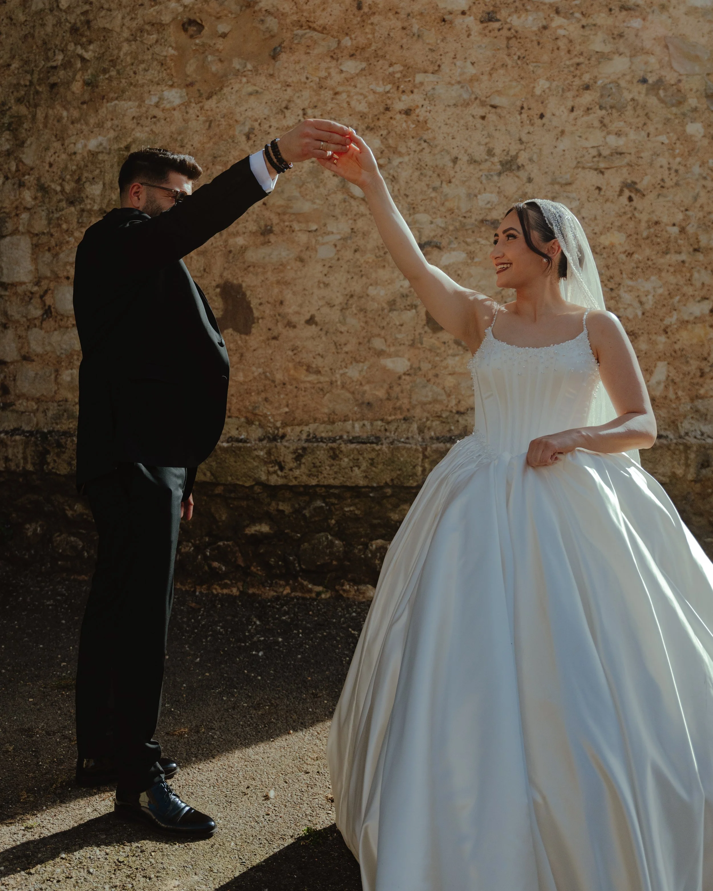 Un couple lors de leur mariage, la femme en robe blanche et le homme en costume noir, dans un décor en pierre, dansant et souriant.