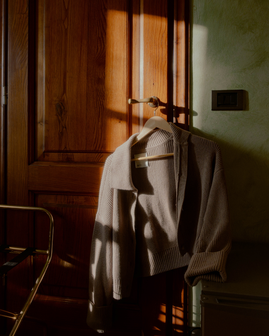 Veste beige suspendue sur un cintre, accrochée à une porte en bois avec de la lumière naturelle qui crée des ombres.