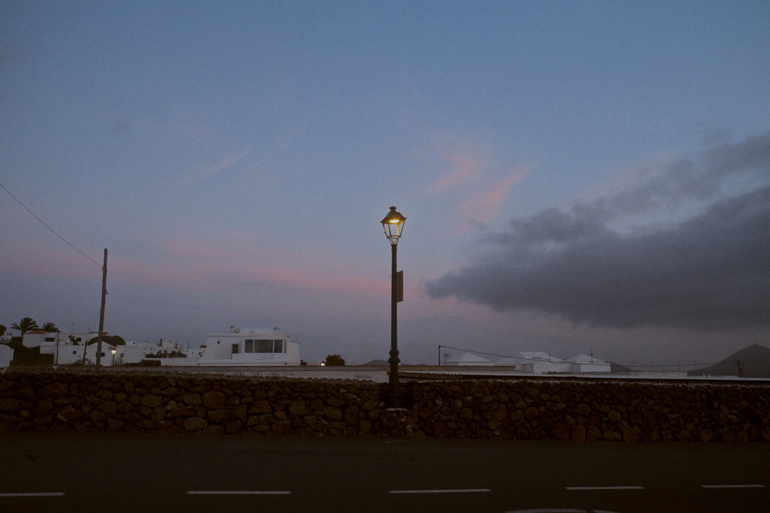 Une rue avec un lampadaire allumé, des maisons blanches, une clôture en pierre et un ciel au crépuscule avec des nuages.