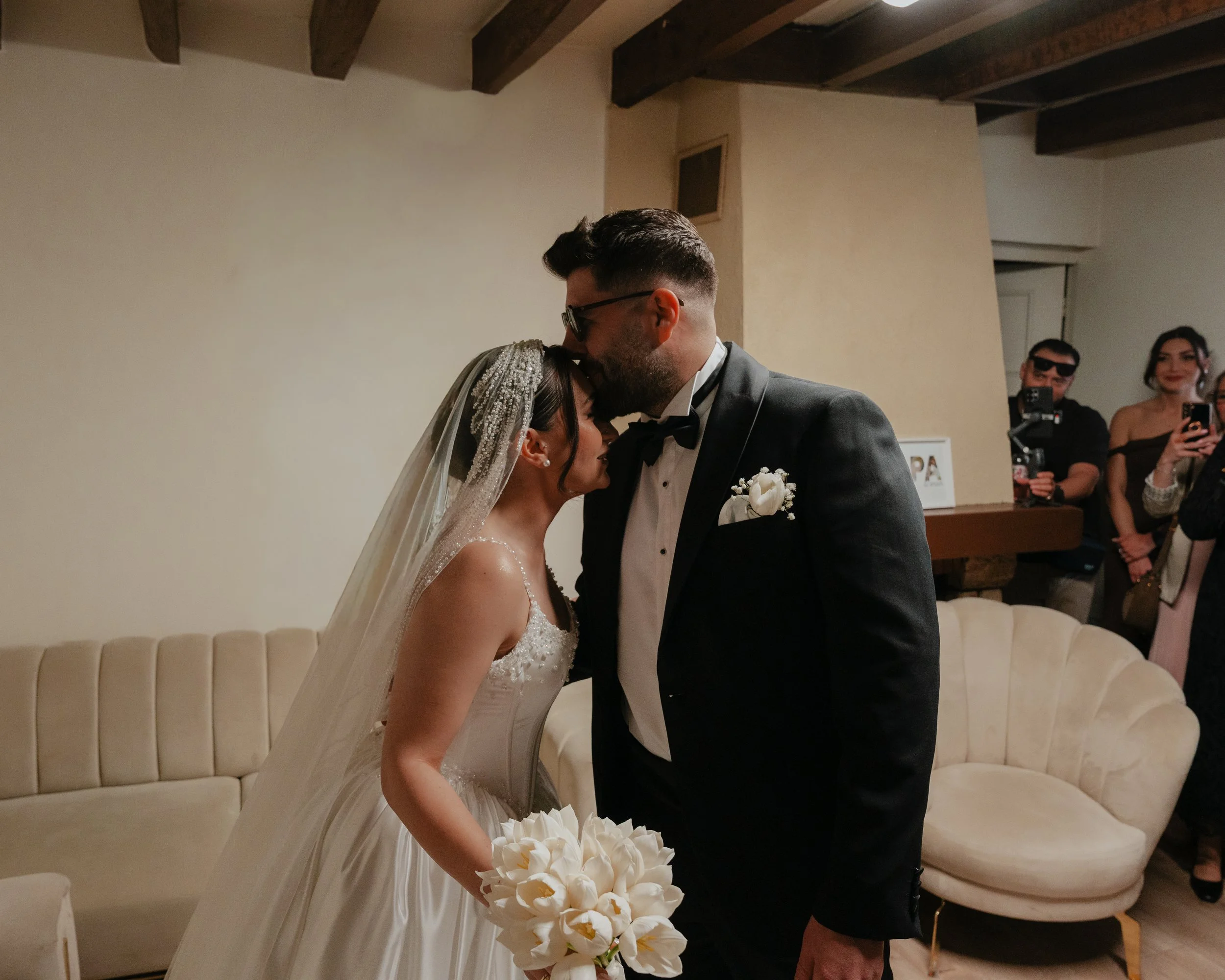 Un couple de mariés lors de leur mariage, échangeant un baiser dans une pièce avec un canapé beige et des invités qui prennent des photos en arrière-plan.