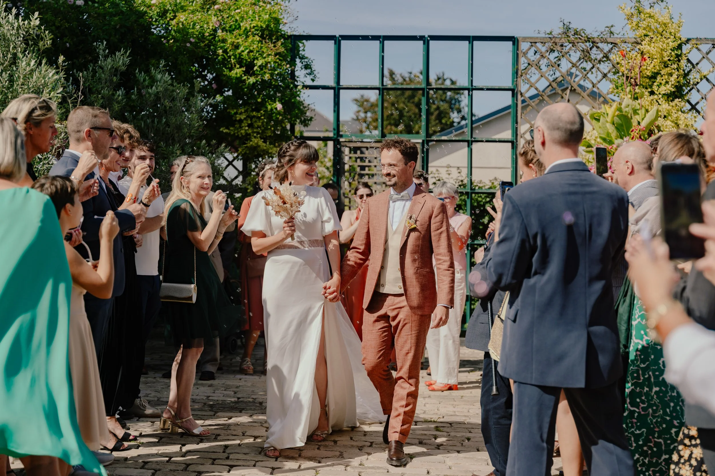 Un couple de mariés marche main dans la main, entouré de leurs invités lors d'une cérémonie en plein air, sous un ciel ensoleillé et entouré de verdure.