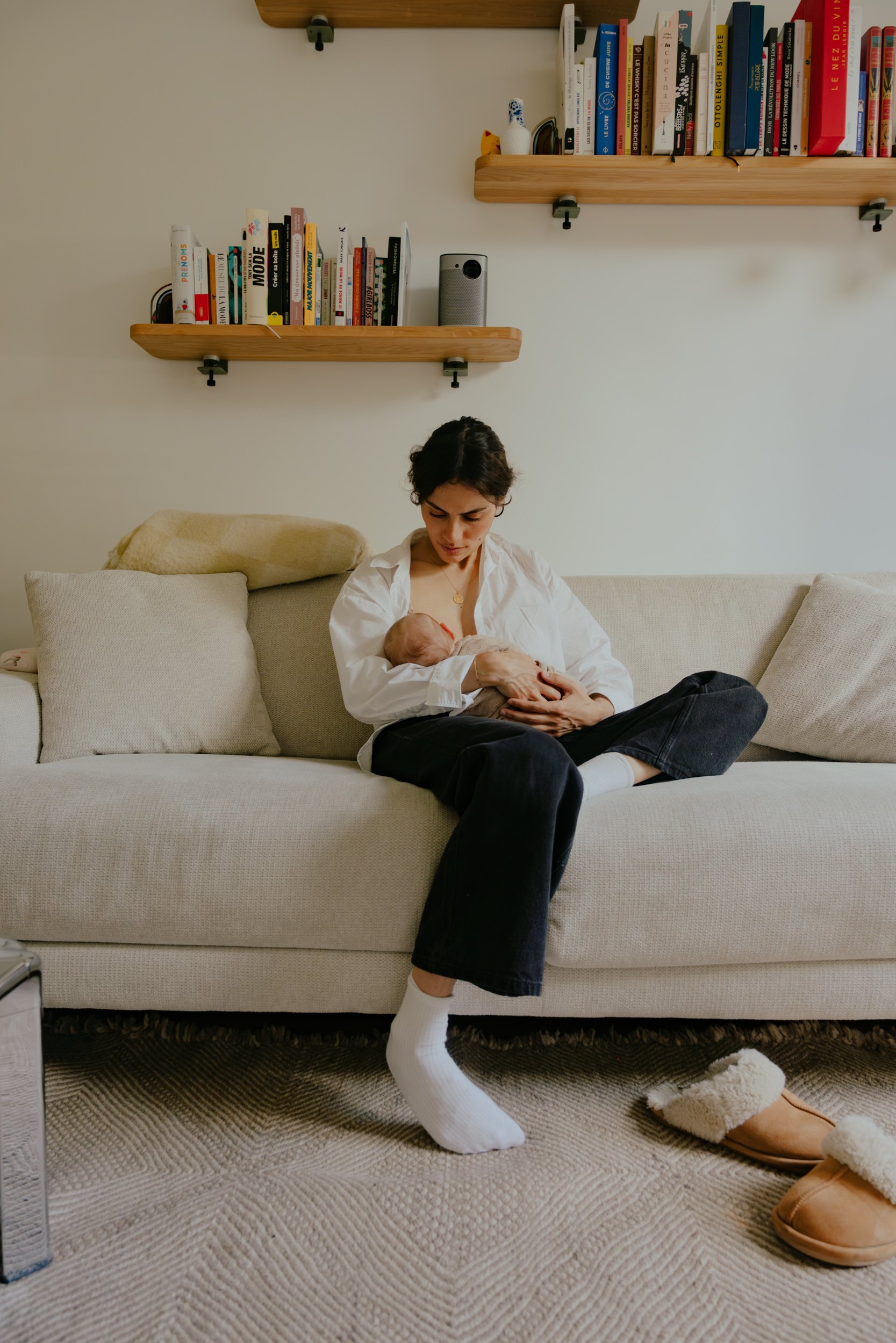 Une femme jeune assise sur un canapé beige, portant des chaussons en peluche et regardant un bébé qu'elle tient dans ses bras. La pièce a deux étagères en bois fixées au mur derrière elle, remplies de livres, une enceinte et un petit objet en forme de chien en peluche sur le sol.