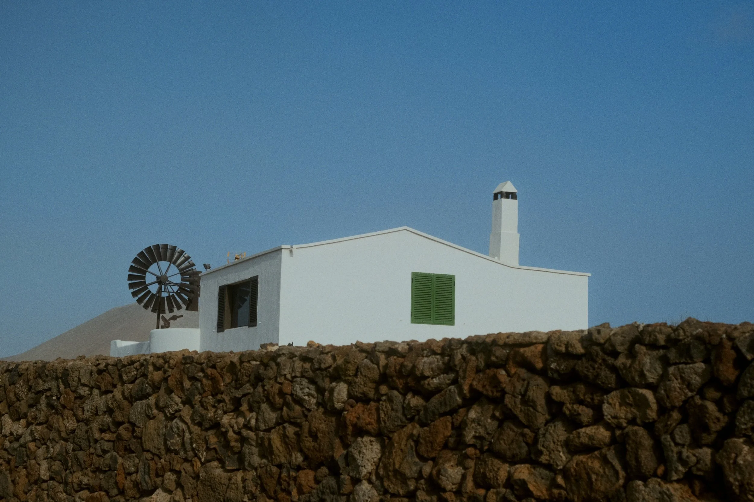 Une maison blanche avec une cheminée et une fenêtre à volets verts, un moulin à vent, et un mur de pierre en premier plan, sous un ciel bleu clair.