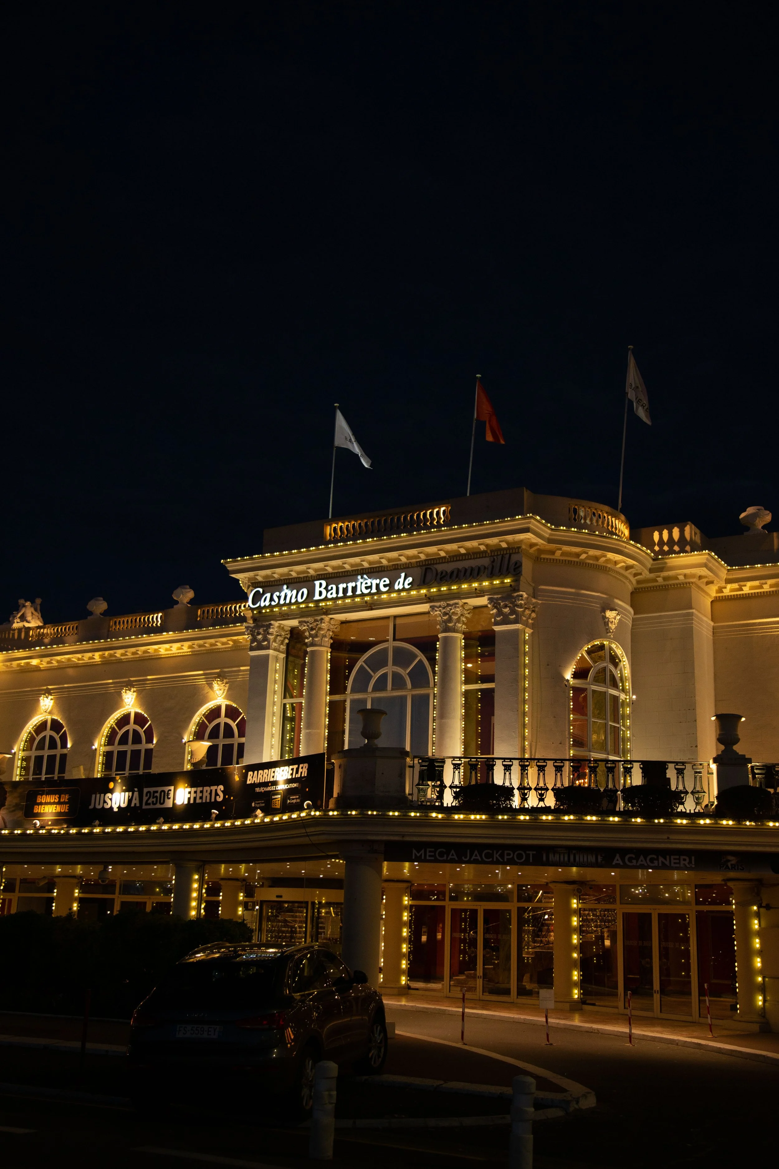 Vue nocturne du bâtiment du Casino Barrière de Deauville illuminé, avec drapeaux flottant au sommet, sur une façade ornée de lumières.