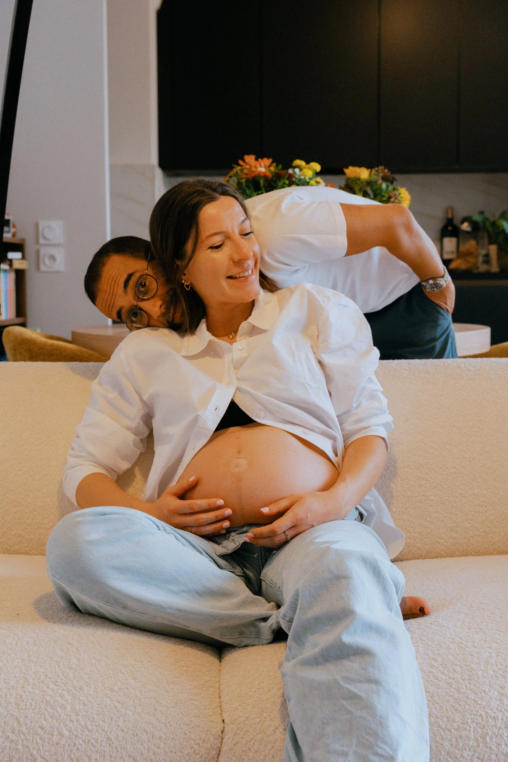 Une femme enceinte souriante assise sur un canapé, une autre personne incline son buste vers elle, dans une maison chaleureuse décorée avec des fleurs et du vin en arrière-plan.