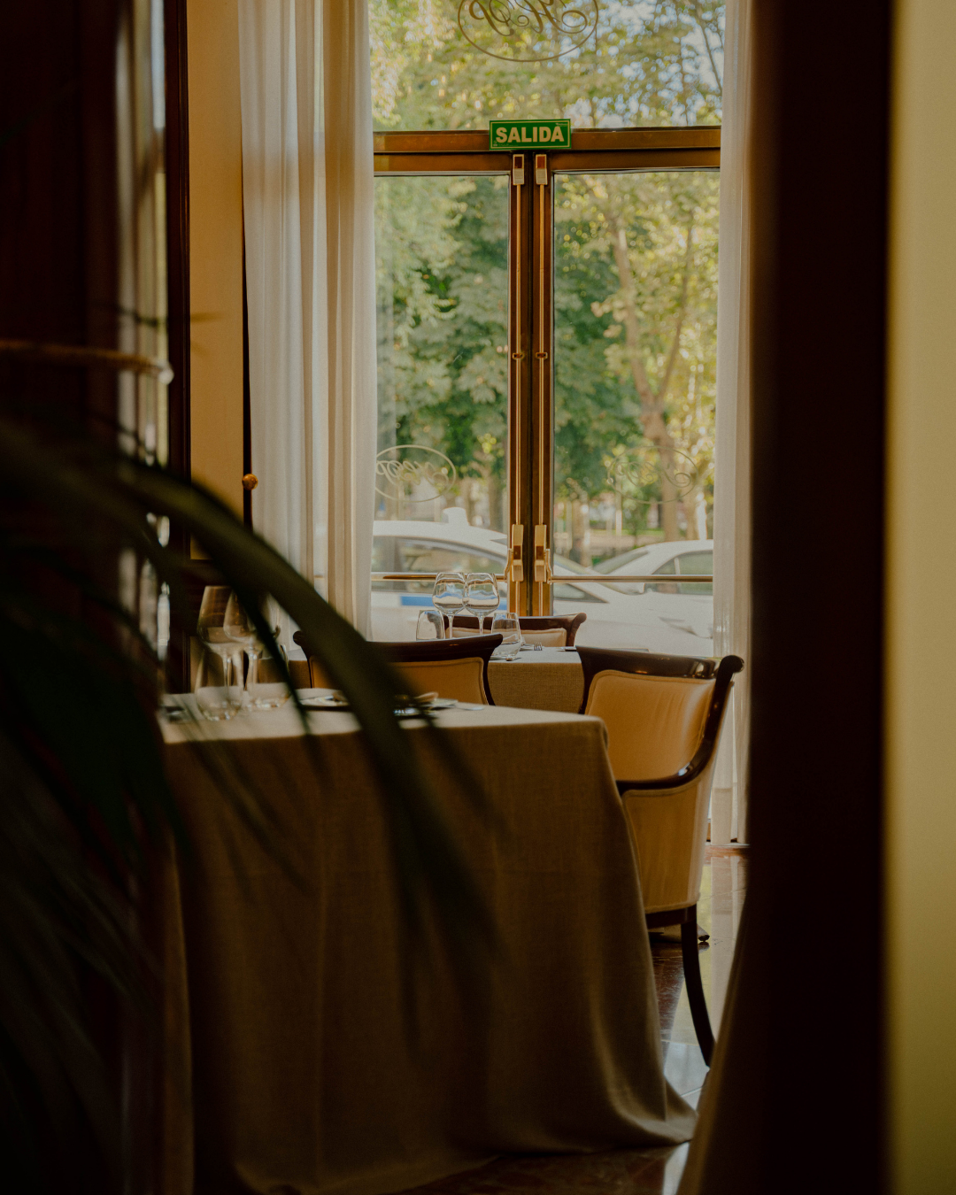 Salle à manger d'un restaurant vue à travers une porte ouverte, avec tables dressées, verres et nappe, et vue sur arbres et voitures à l'extérieur.