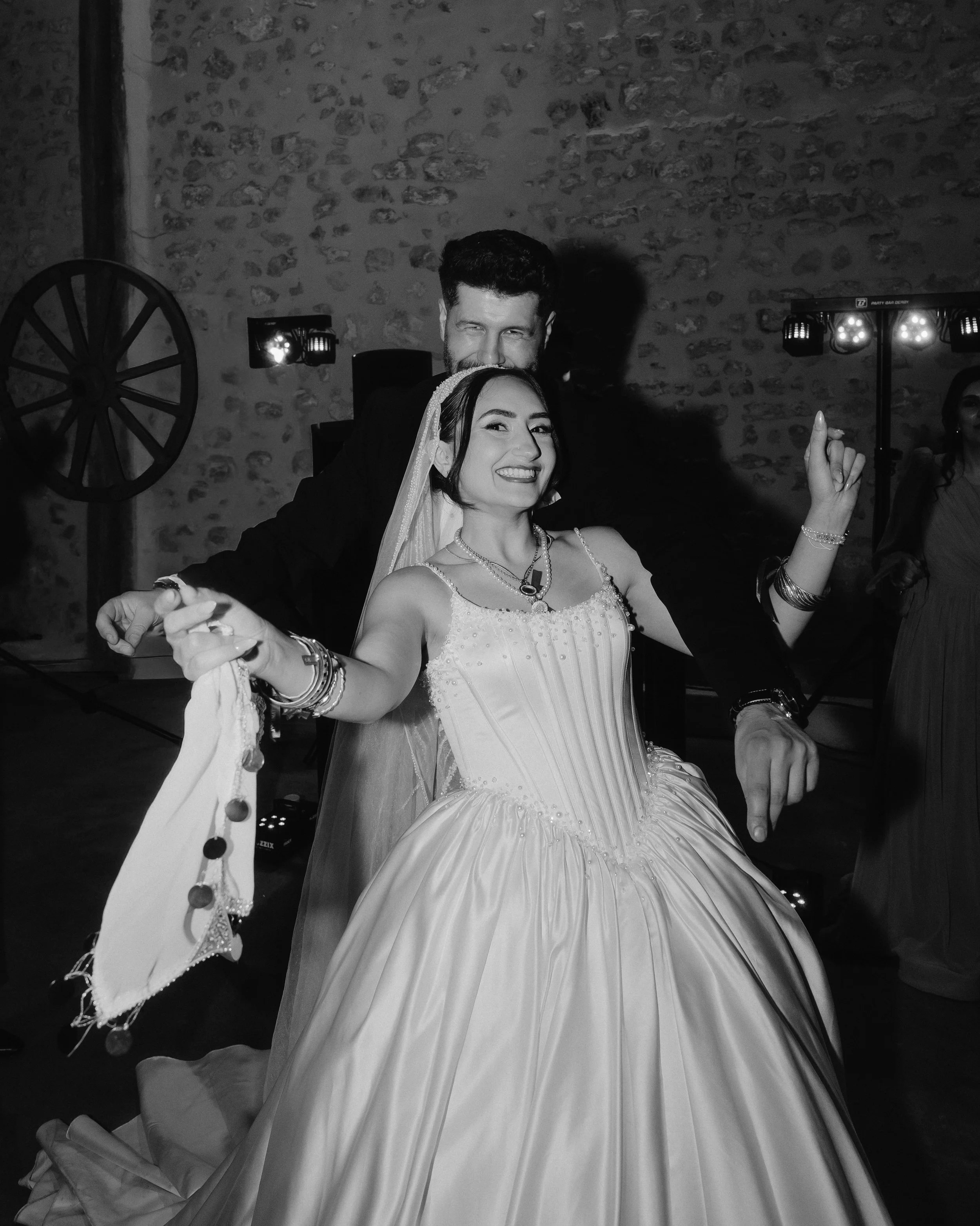 Un couple danse lors d'une réception, la femme porte une robe de mariée et le homme un costume. La scène est en noir et blanc. La femme affiche un sourire large et porte des bijoux, tandis que l'homme se trouve derrière elle, avec un regard direction