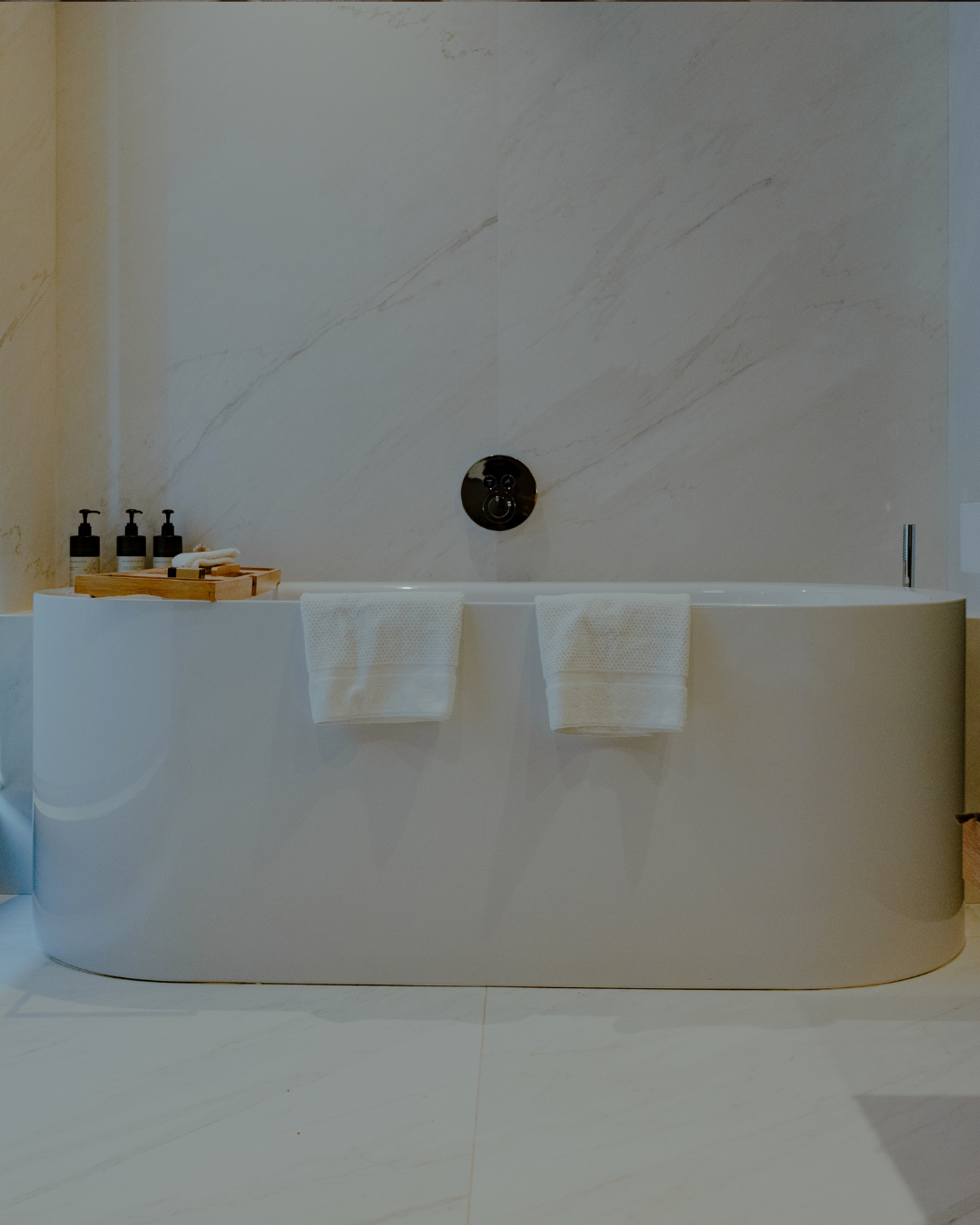Baignoire moderne dans une salle de bain élégante, avec deux serviettes blanches suspendues et des bouteilles de produits de bain sur un plateau en bois.