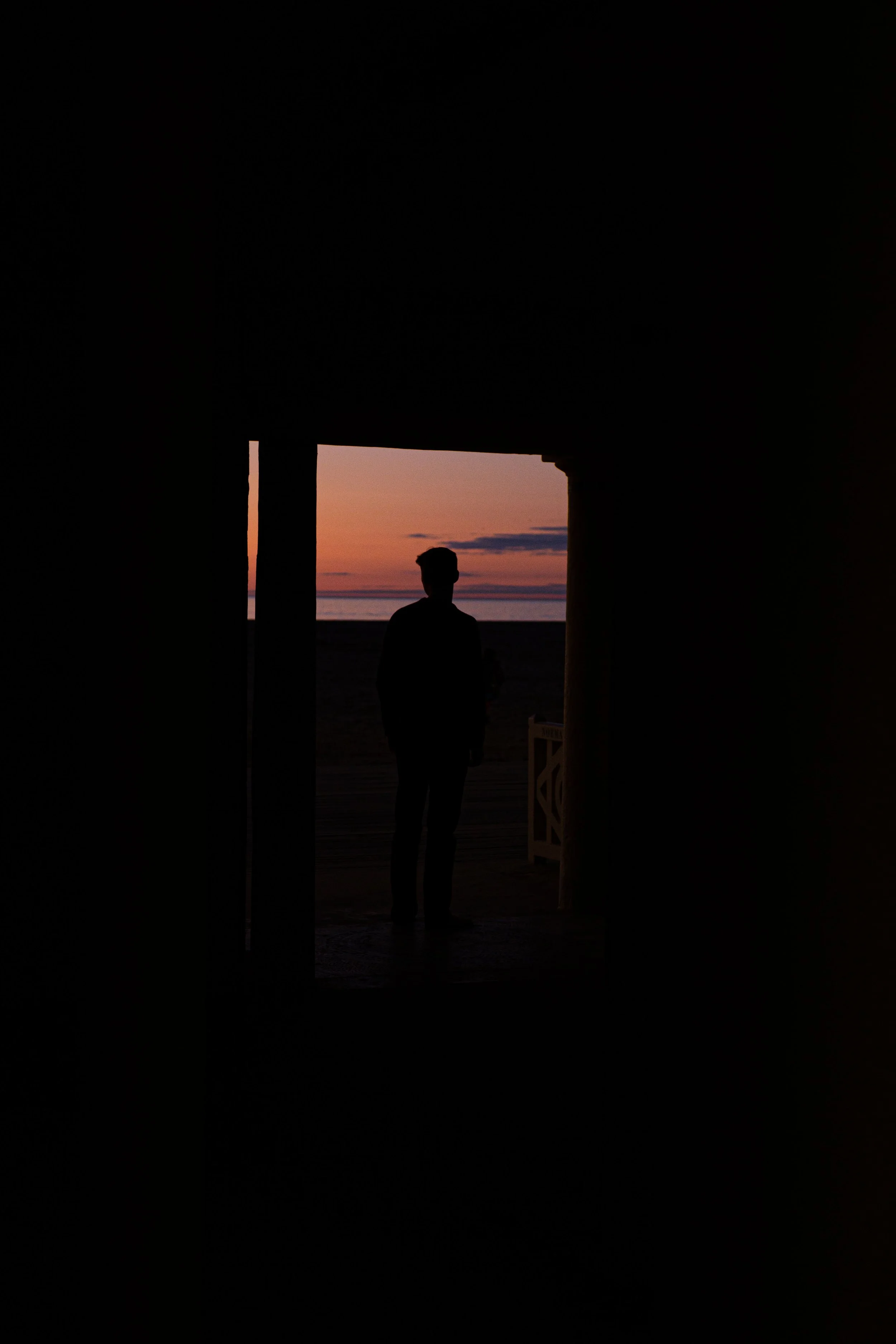 Silhouette d'une personne debout devant un coucher de soleil sur la plage, vue depuis l'intérieur d'une structure sombre.