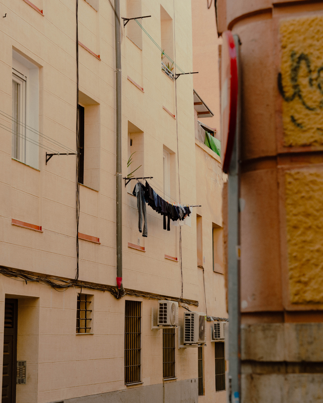Vue d'un immeuble résidentiel avec des fenêtres, des climatiseurs, des lignes de lavage avec des vêtements suspendus, et un mur de brique à droite.