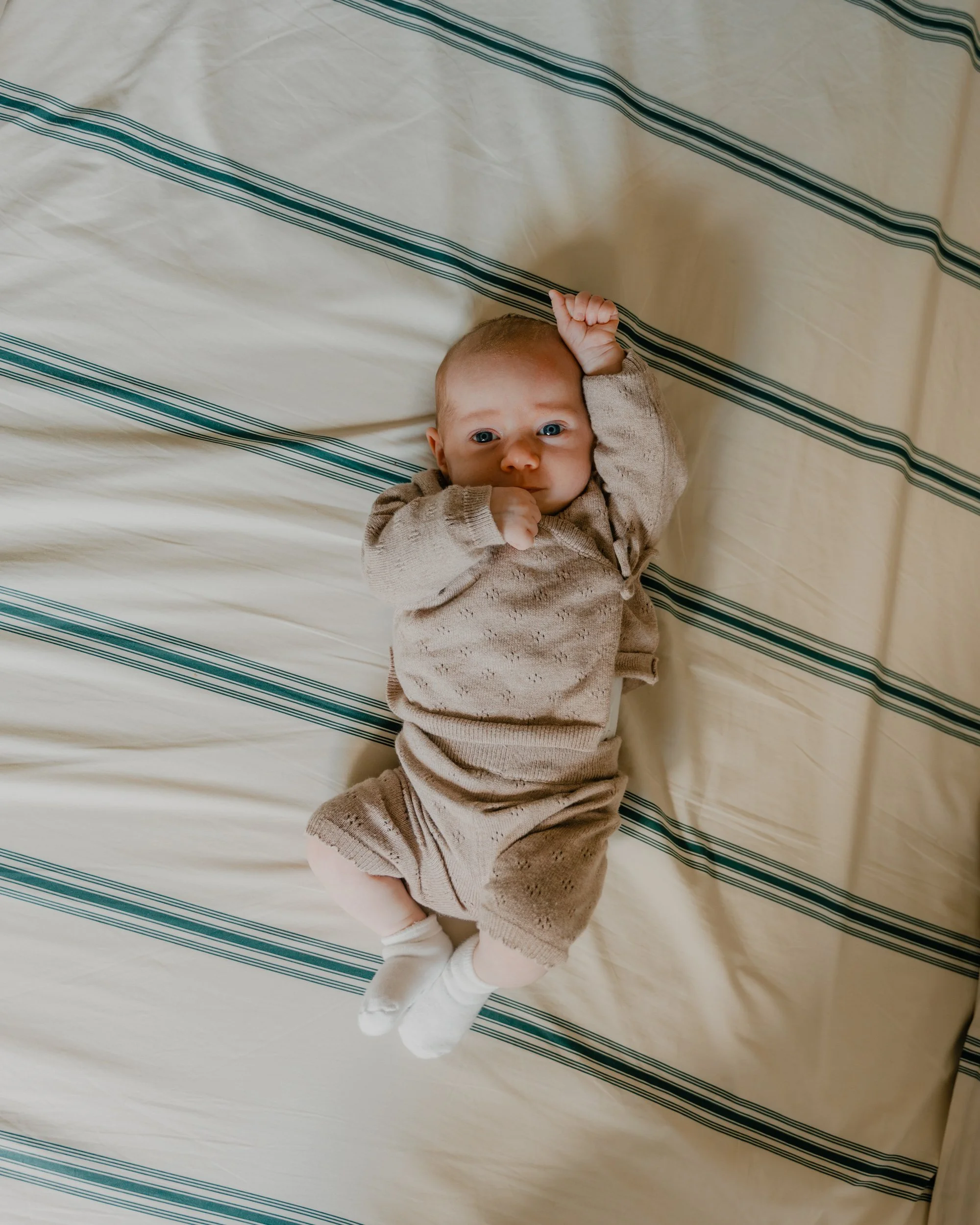 Un bébé allongé sur un lit, regardant l'objectif, avec une main près de sa tête, portant un pyjama beige et des chaussettes blanches.