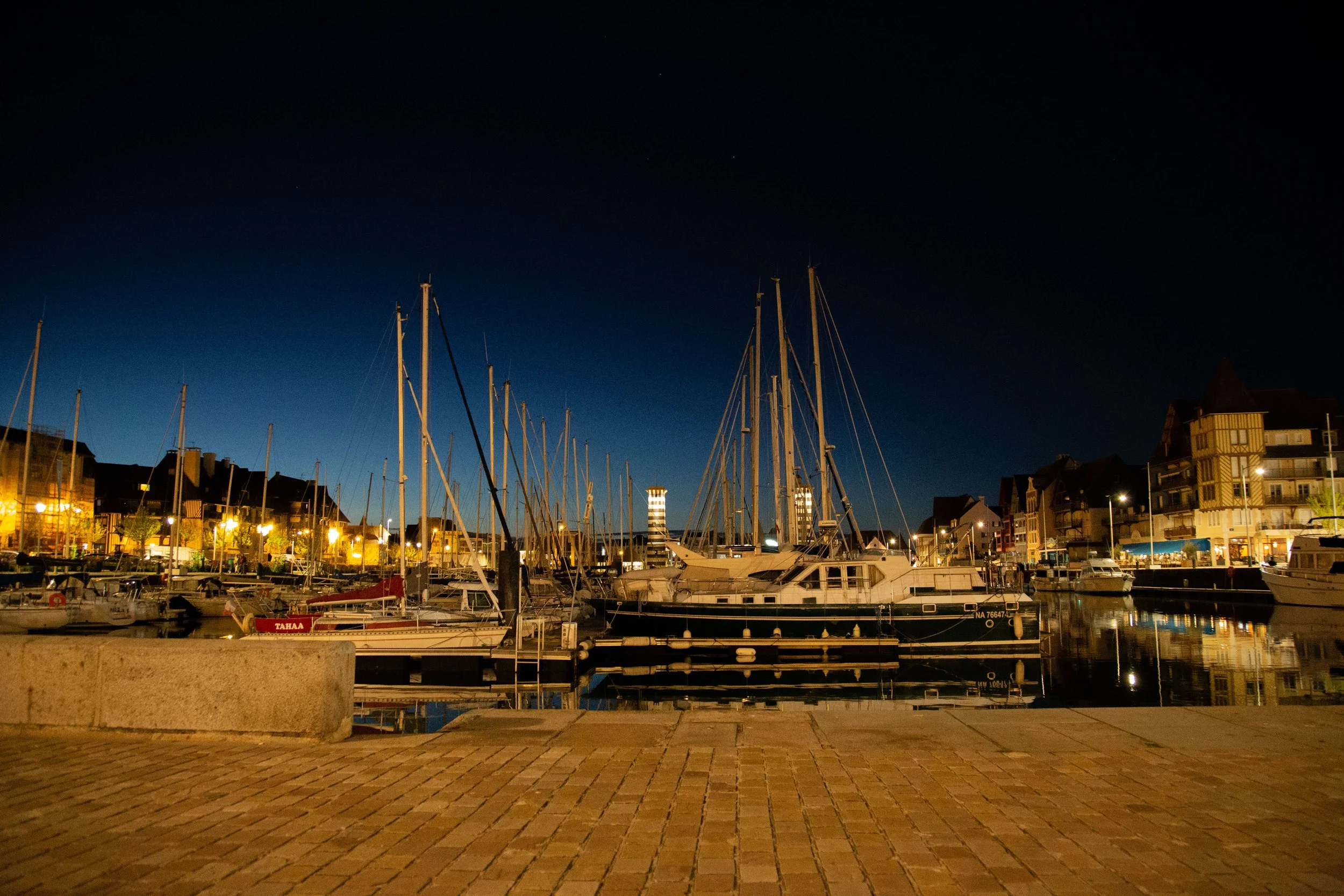 Un port de plaisance avec des yachts et voiliers, bâtiments résidentiels en arrière-plan, illuminés la nuit, reflet de l'eau calme.