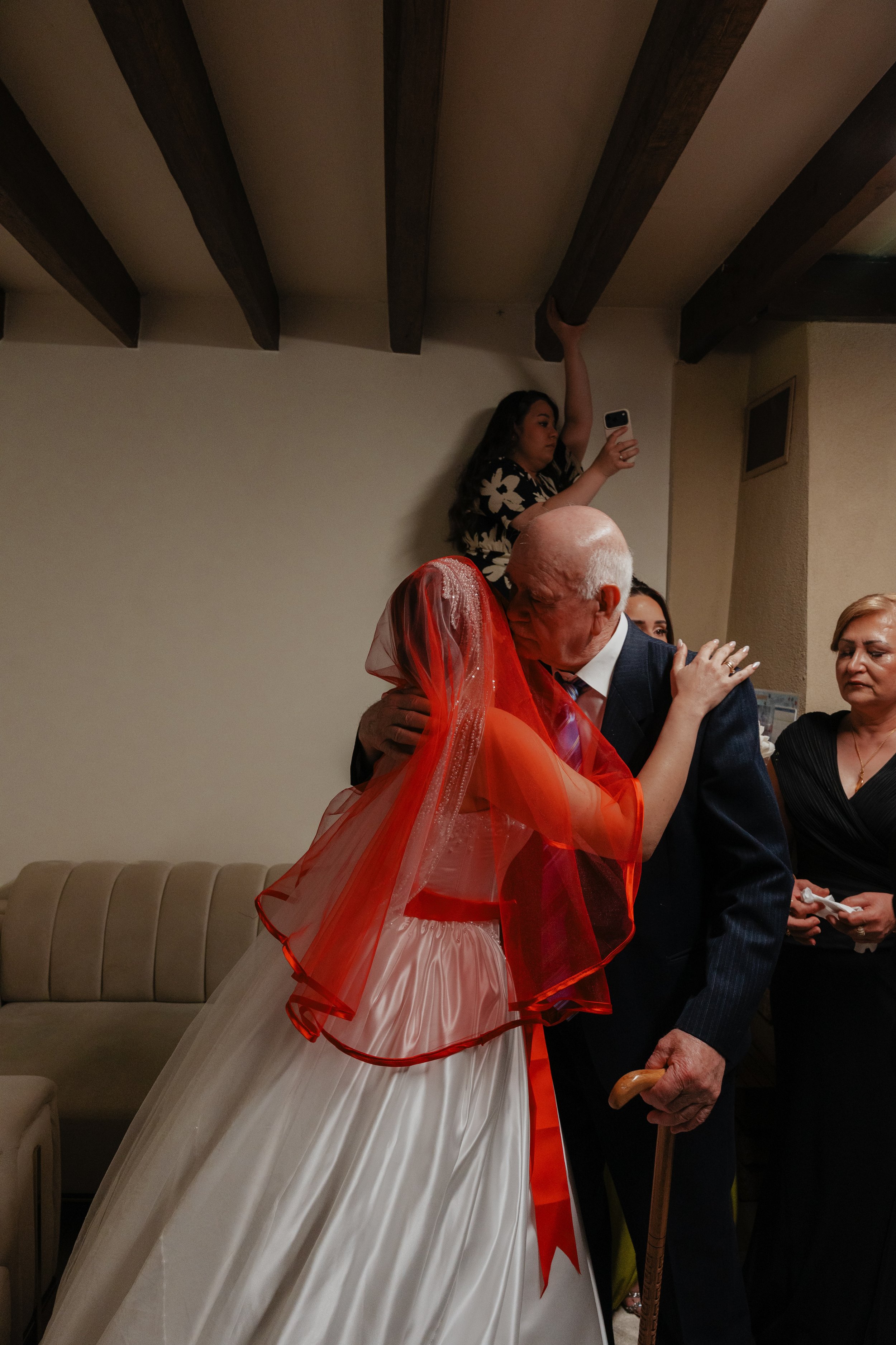 Une femme en robe de mariage avec un voile orange et une autre personne embrassent lors d'une célébration. Une femme en haut prend une photo avec son téléphone, dans un intérieur avec plafond en poutres en bois.