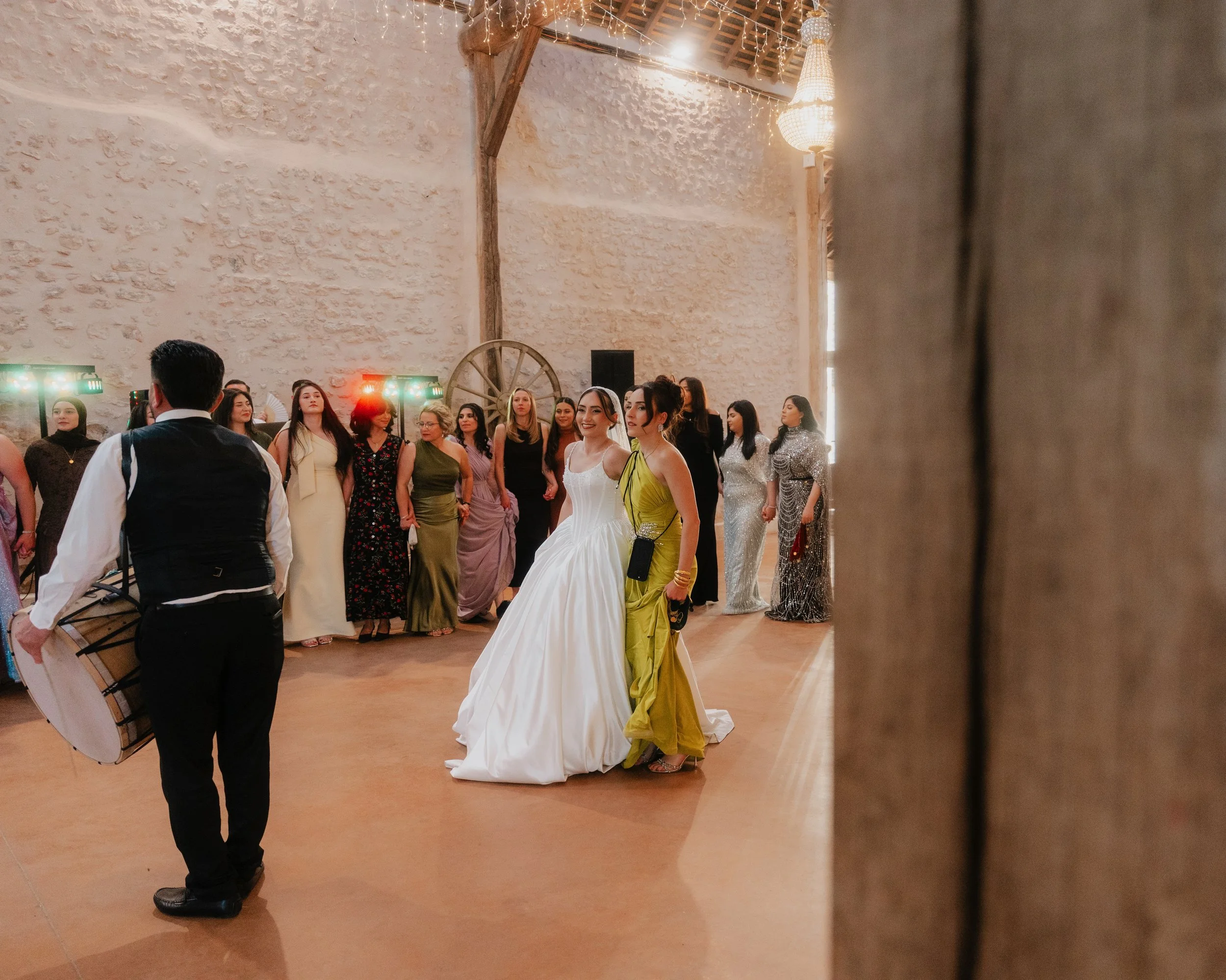 Groupe de personnes lors d'une cérémonie ou d'une fête dans une grande salle avec murs en pierre, éclairage chaud et décoration lumineuse suspendue, plusieurs femmes habillées en robes élégantes, deux femmes au centre souriantes, un homme en veston d