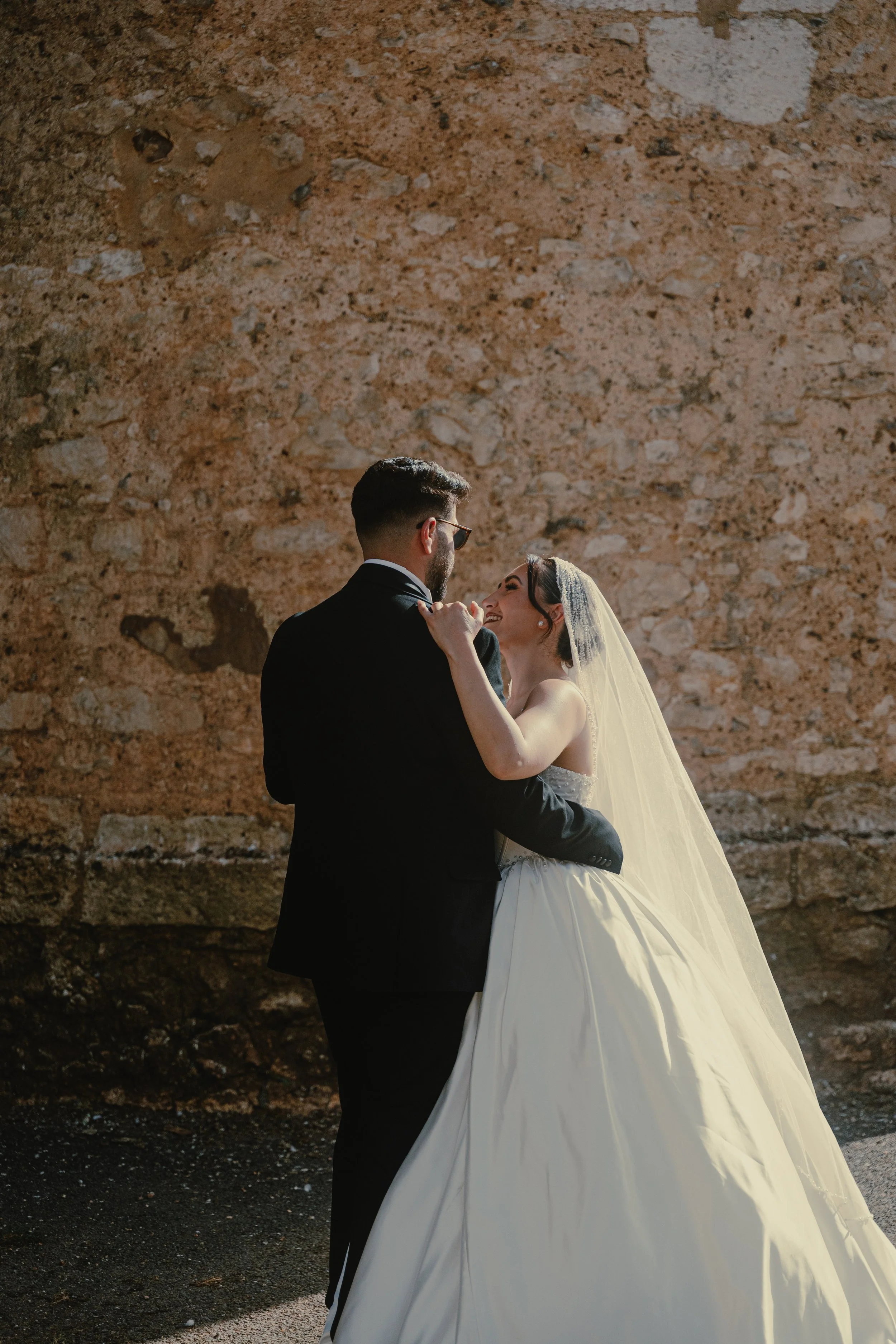 Un couple marié danse près d'un mur en pierre, la femme porte une robe de mariage blanche et un voile, et l'homme porte un costume noir.