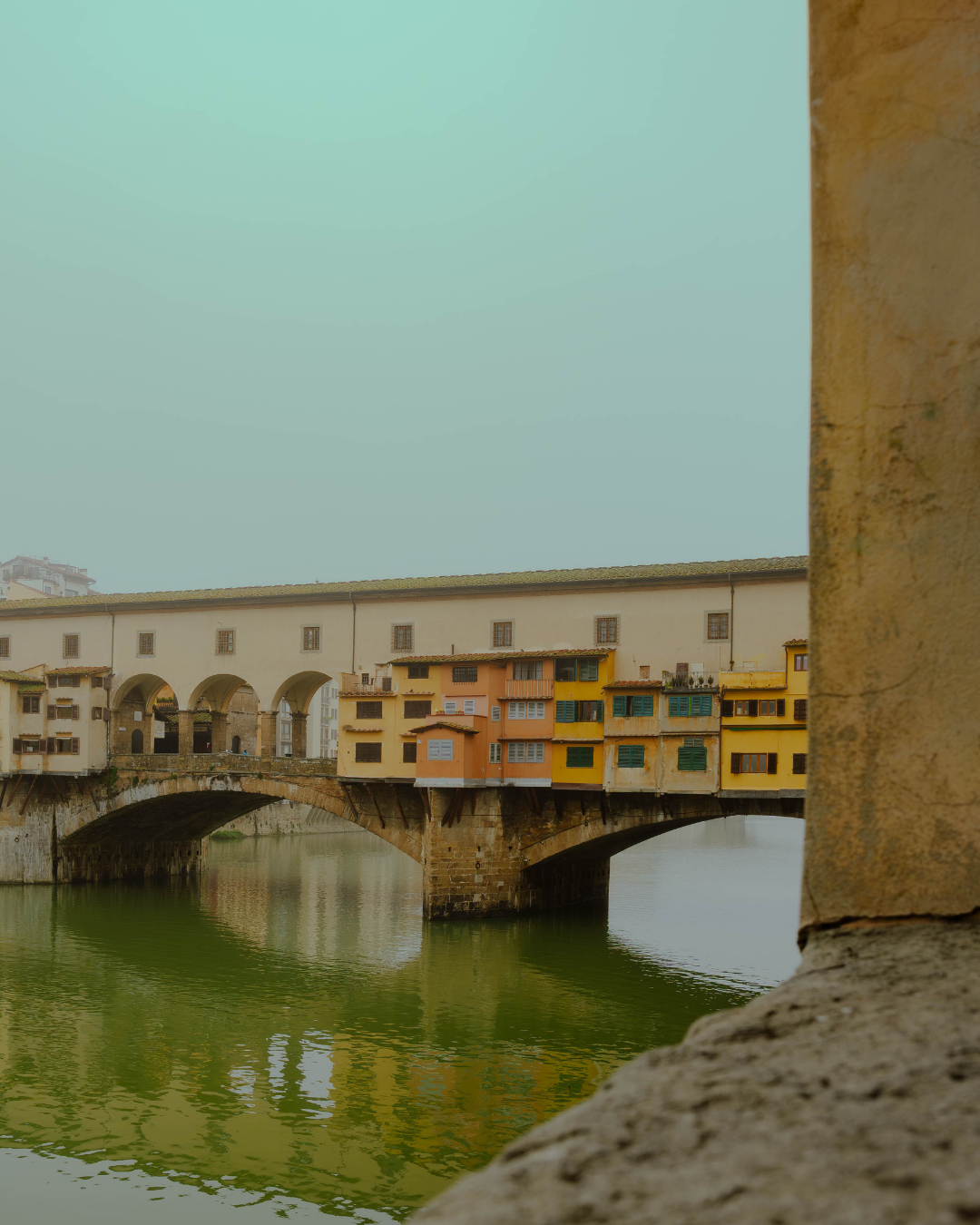 Vue d'un vieux pont avec des maisons colorées en dessous, reflet dans l'eau. Part d'une colonne en pierre à droite.