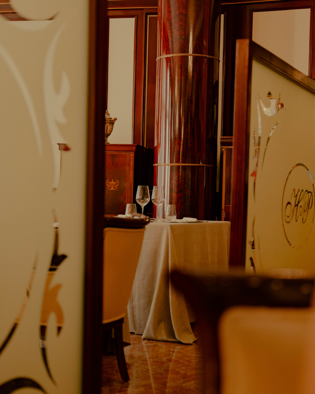 Salle à manger élégante dans un restaurant, vue partielle à travers une porte ornée de motifs dorés, table avec nappe beige, trois verres à vin, ambiance chaleureuse et sophistiquée.