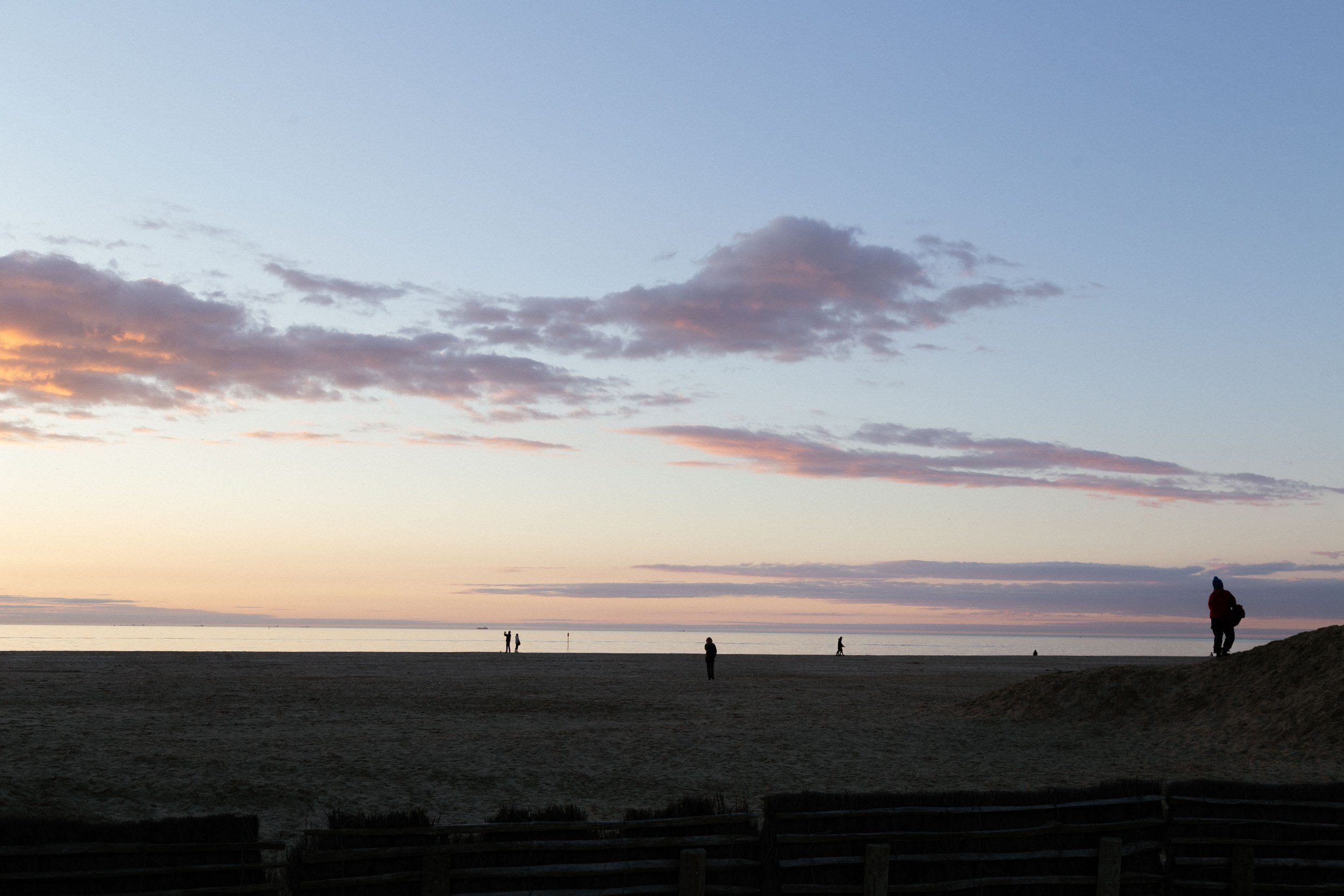 Vue d'une plage au coucher du soleil avec le ciel coloré, quelques nuages, et plusieurs personnes marchant ou se tenant au bord de l'eau, dont une personne en silhouette portant un manteau rouge à droite.
