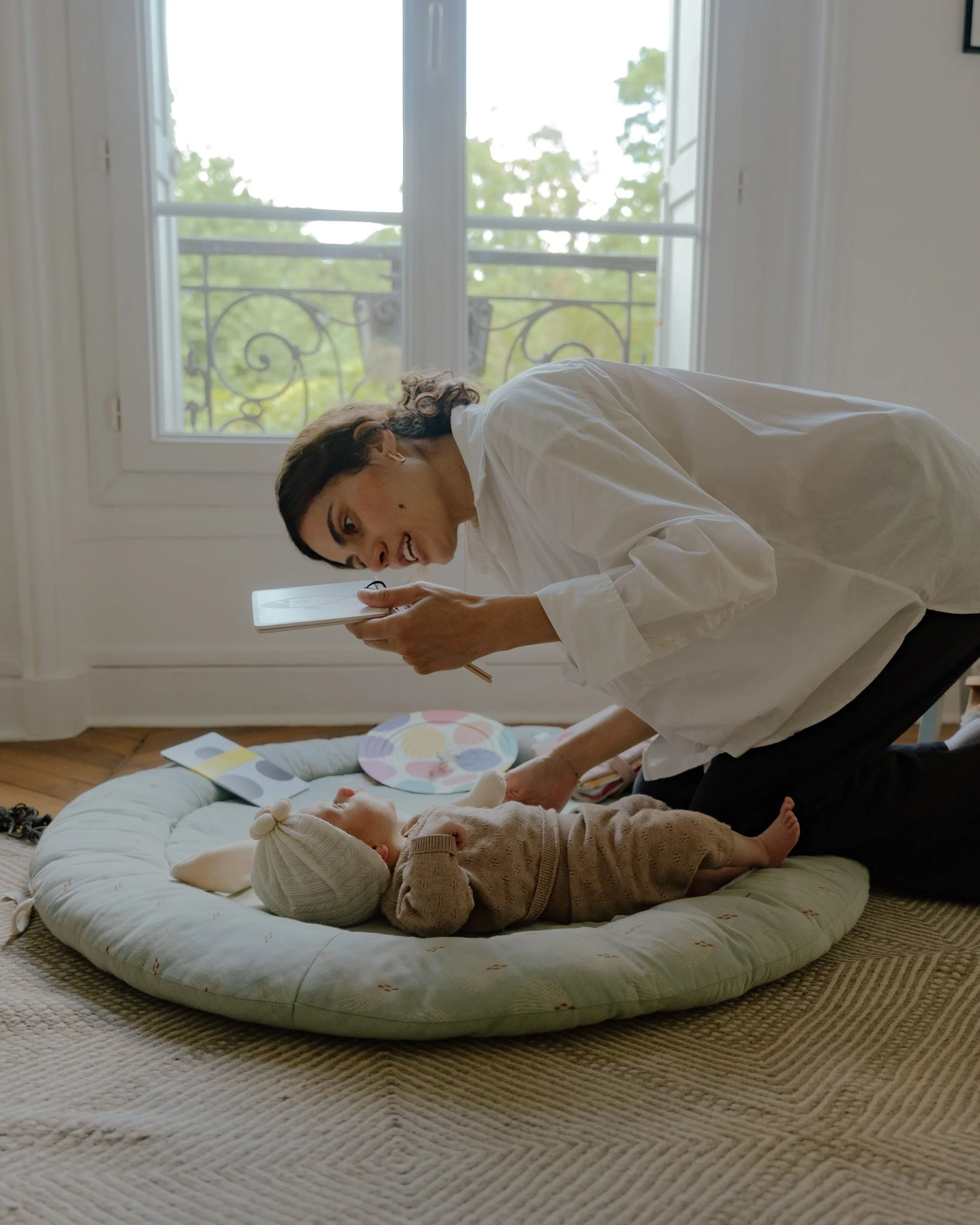 Une femme souriante en chemise blanche observe un bébé allongé sur un tapis de jeu en forme de cercle au sol d'une pièce lumineuse, avec une grande fenêtre en arrière-plan et des décorations colorées autour.