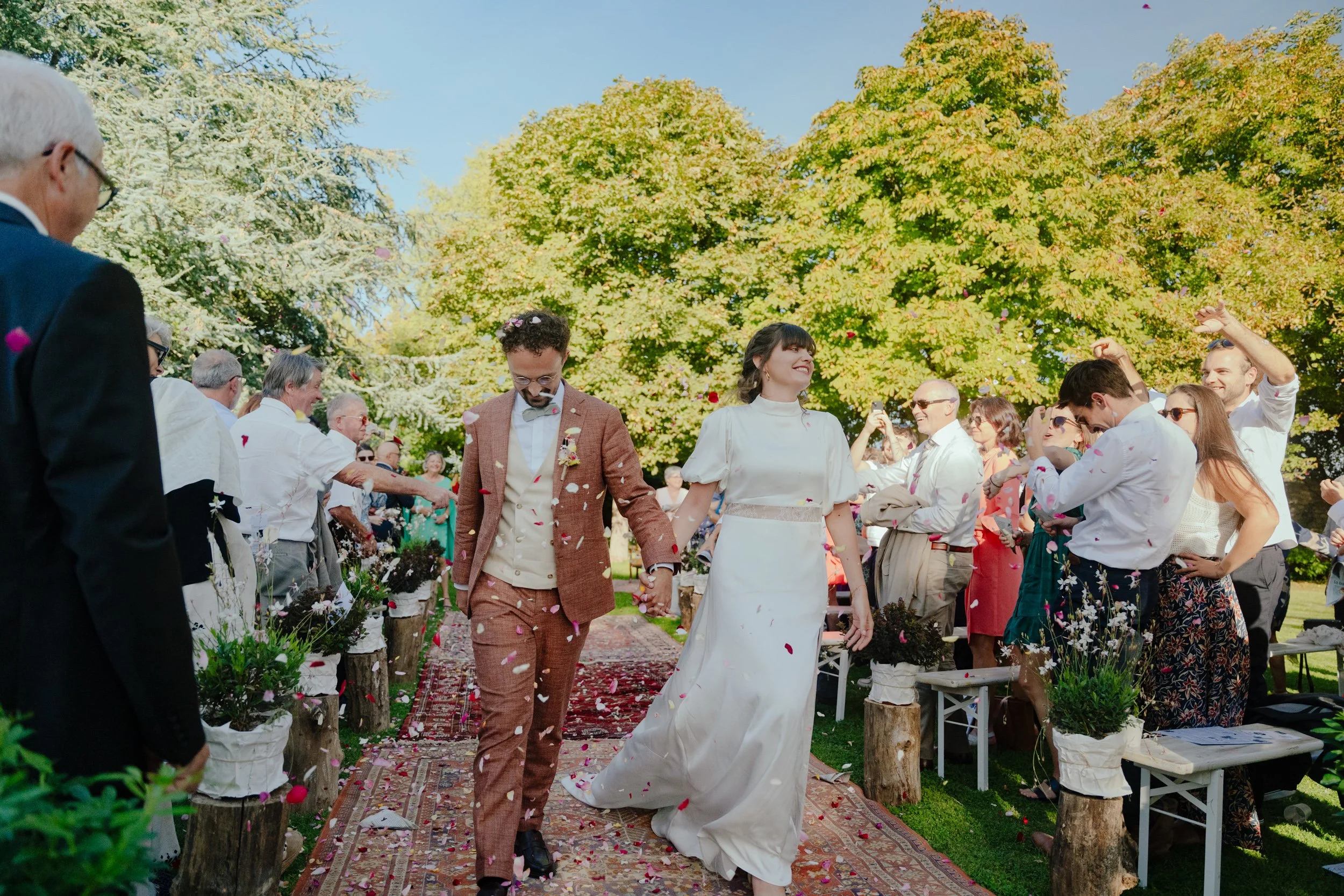 Un couple de mariés marchant main dans la main lors de leur mariage en plein air, entourés par des invités et de grands arbres verts, sous un ciel bleu.