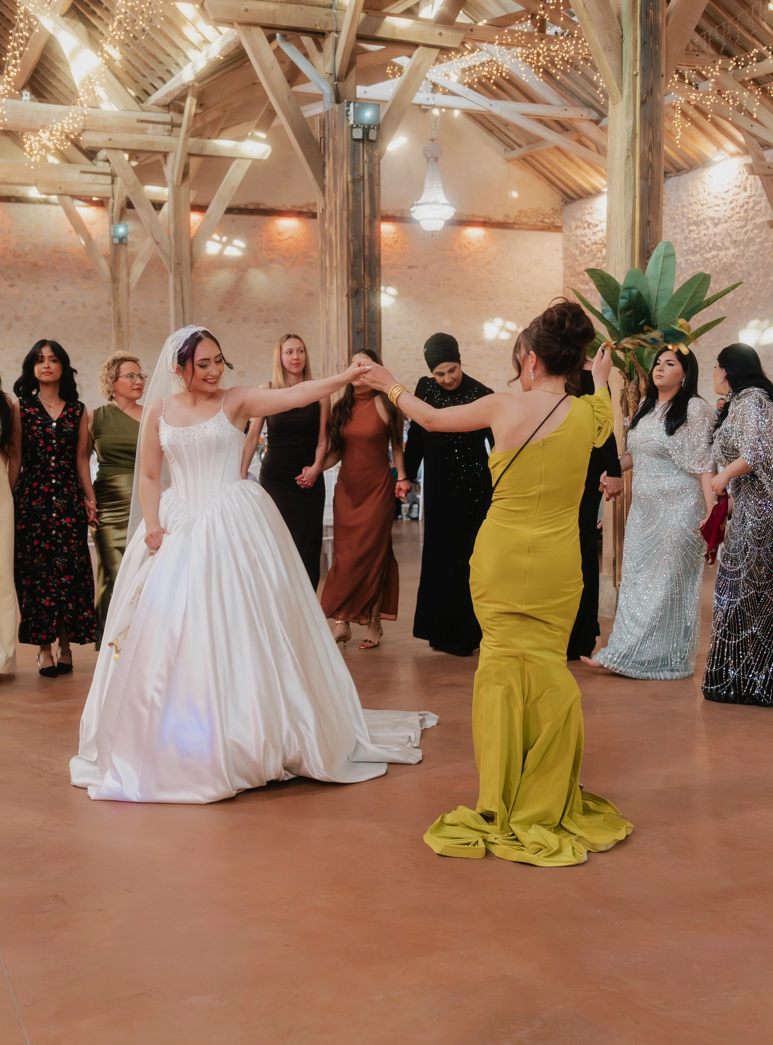 Une mariée en robe blanche danse avec une femme en robe jaune lors d'une célébration dans une salle aux murs en pierre. D'autres personnes en robes élégantes regardent la scène tandis qu'une décoration lumineuse suspendue orne le plafond en bois.