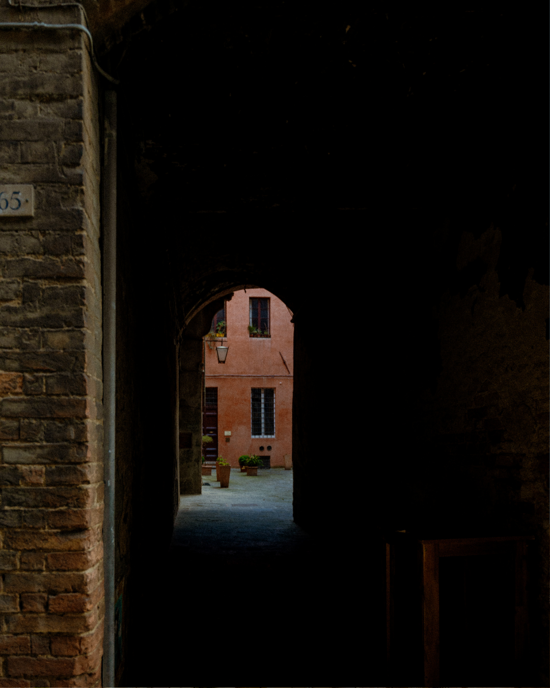 Vue d'une ruelle sombre menant à une place ensoleillée avec un bâtiment rose et des pots de fleurs.