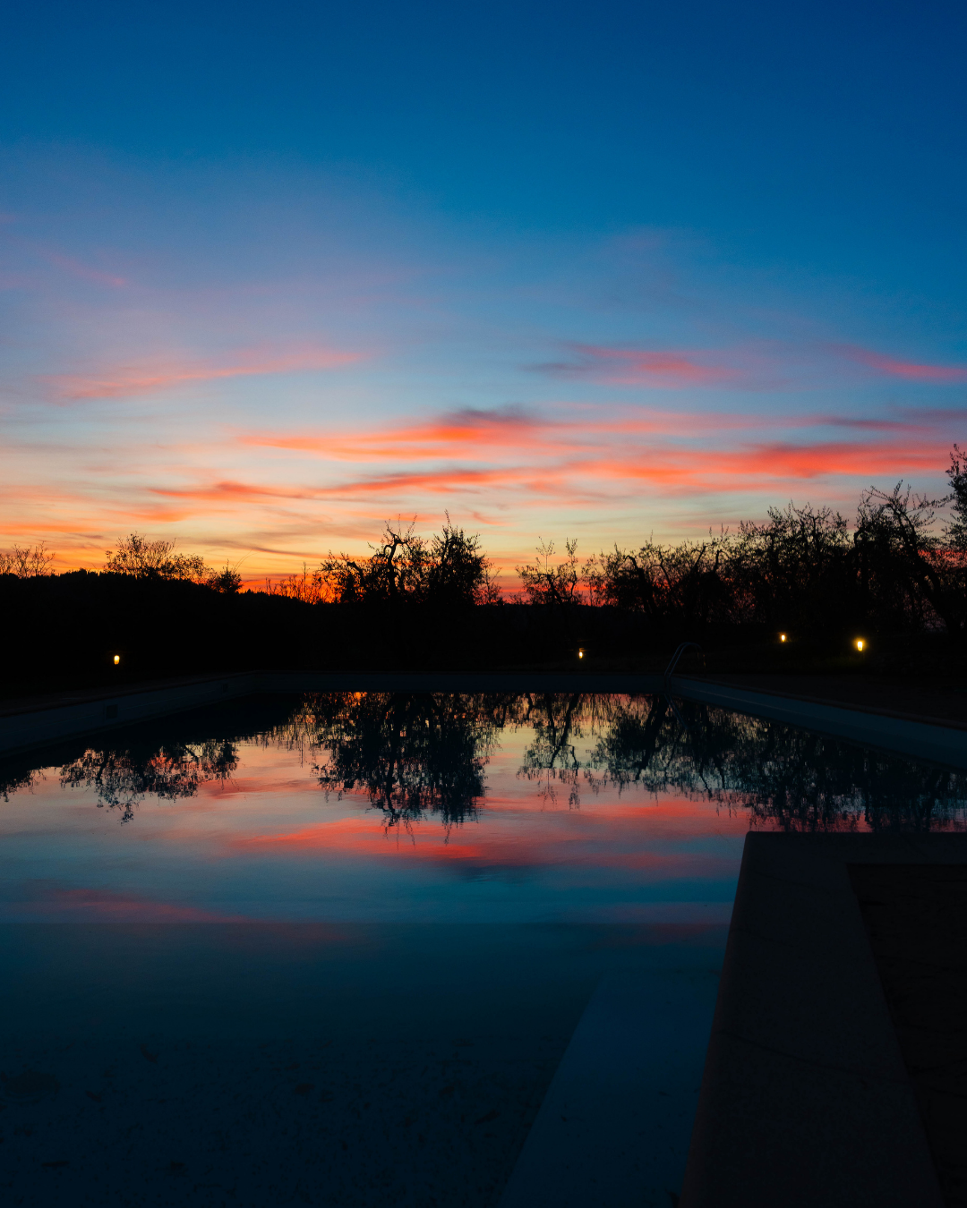 Coucher de soleil avec ciel bleu et nuages roses, reflet dans une piscine, silhouettes d'arbres et petite lumière au bord de la piscine.