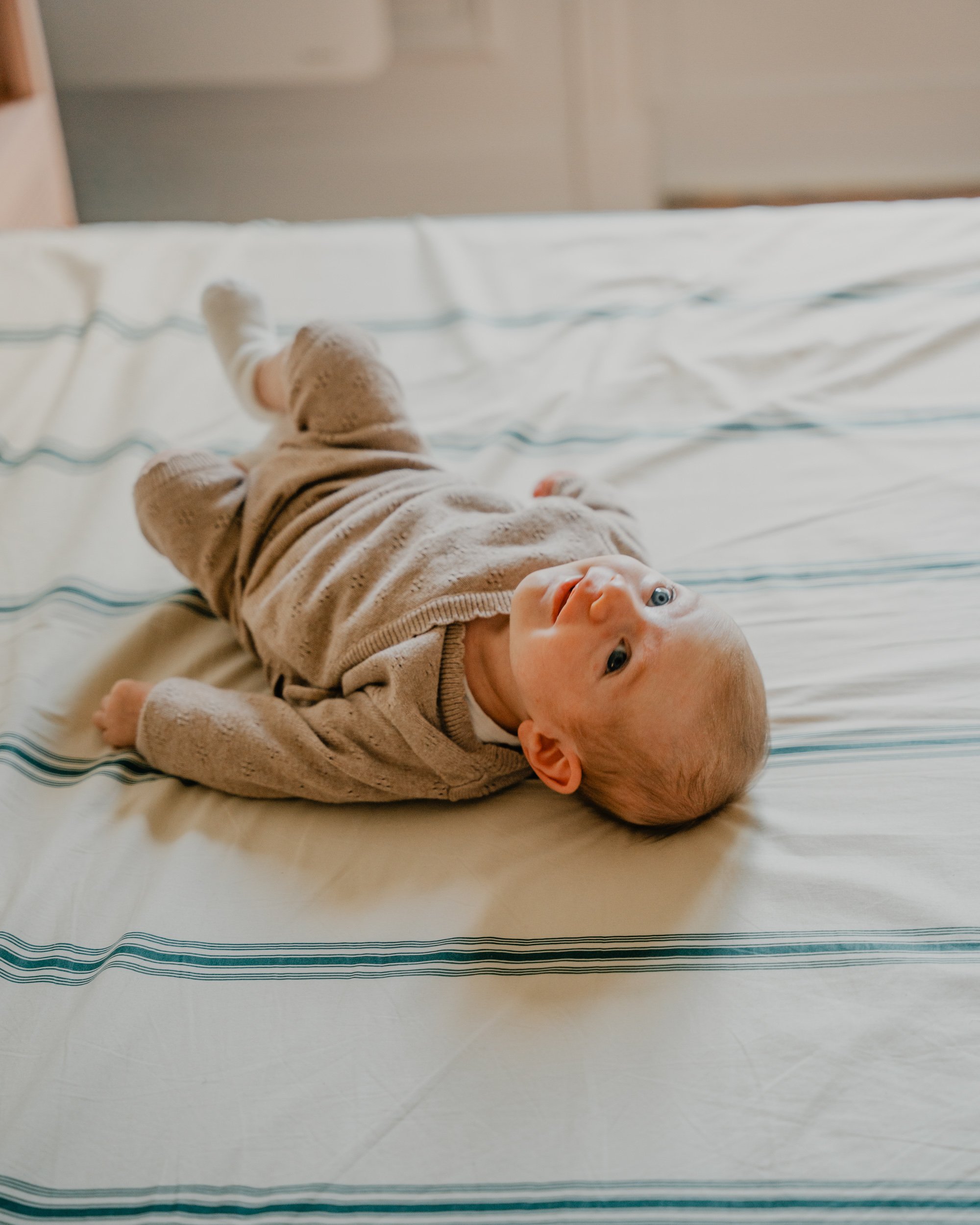 Un bébé allongé sur un lit avec une chemise beige, regardant vers l'appareil photo, avec un lit blanc rayé bleu et blanc.