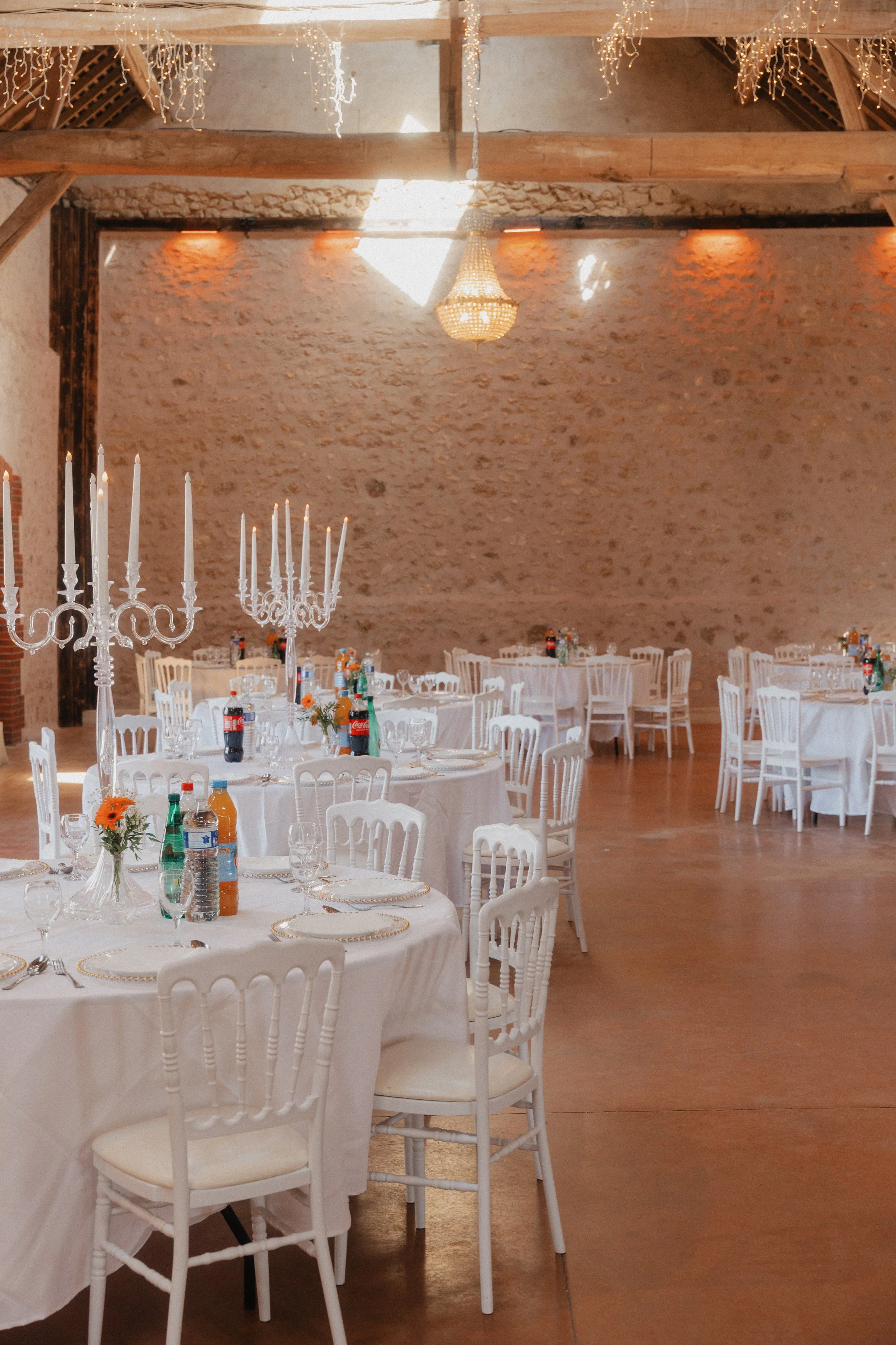 Salle de réception décorée pour un événement, avec des chaises blanches autour de tables rondes, des candélabres blancs, des bouteilles de boisson et des bouquet de fleurs. Mur en pierre et éclairage chaleureux.