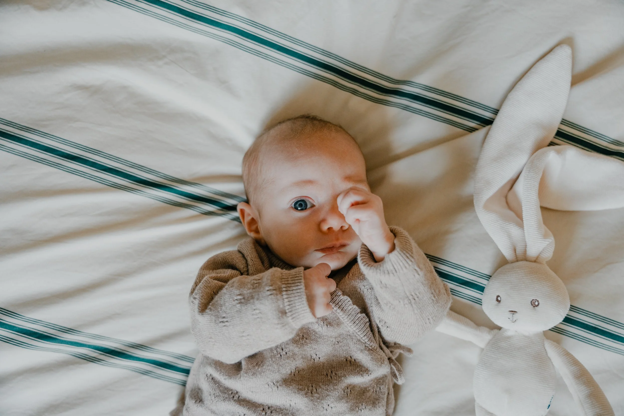 Un bébé allongé sur une couverture ou drap blanc avec des rayures bleues, faisant un geste avec la main et regardant l'appareil photo.