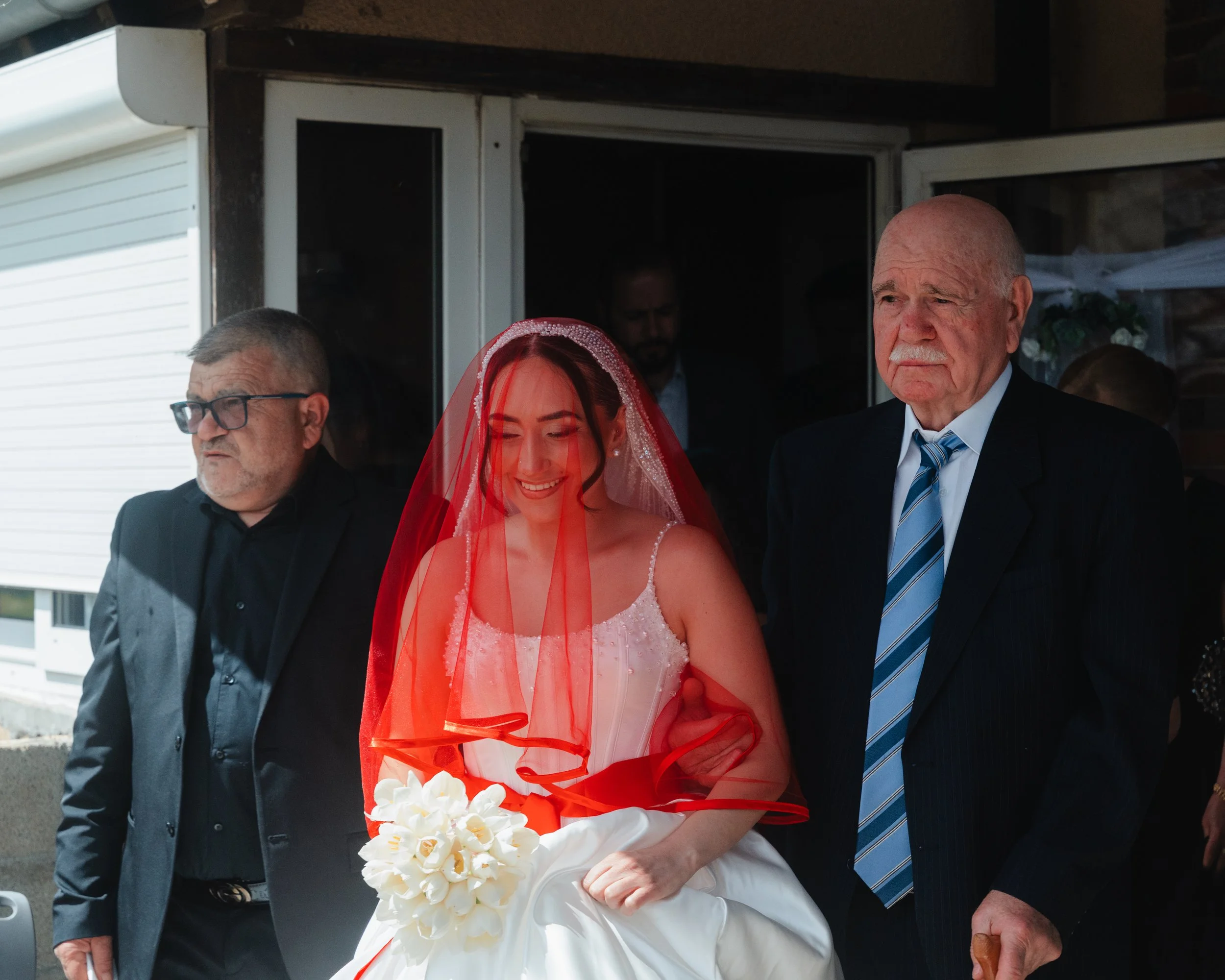 Une femme en robe blanche de mariage avec un voile rouge tient un bouquet de fleurs blanches, entourée de trois hommes en costume lors d'une cérémonie ou d'une fête
