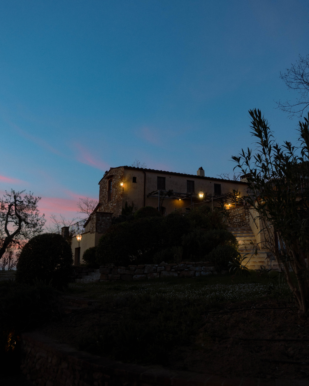 Une maison ancienne en pierre sur une colline, éclairée par des lampes, avec un ciel en soirée ou à l'aube