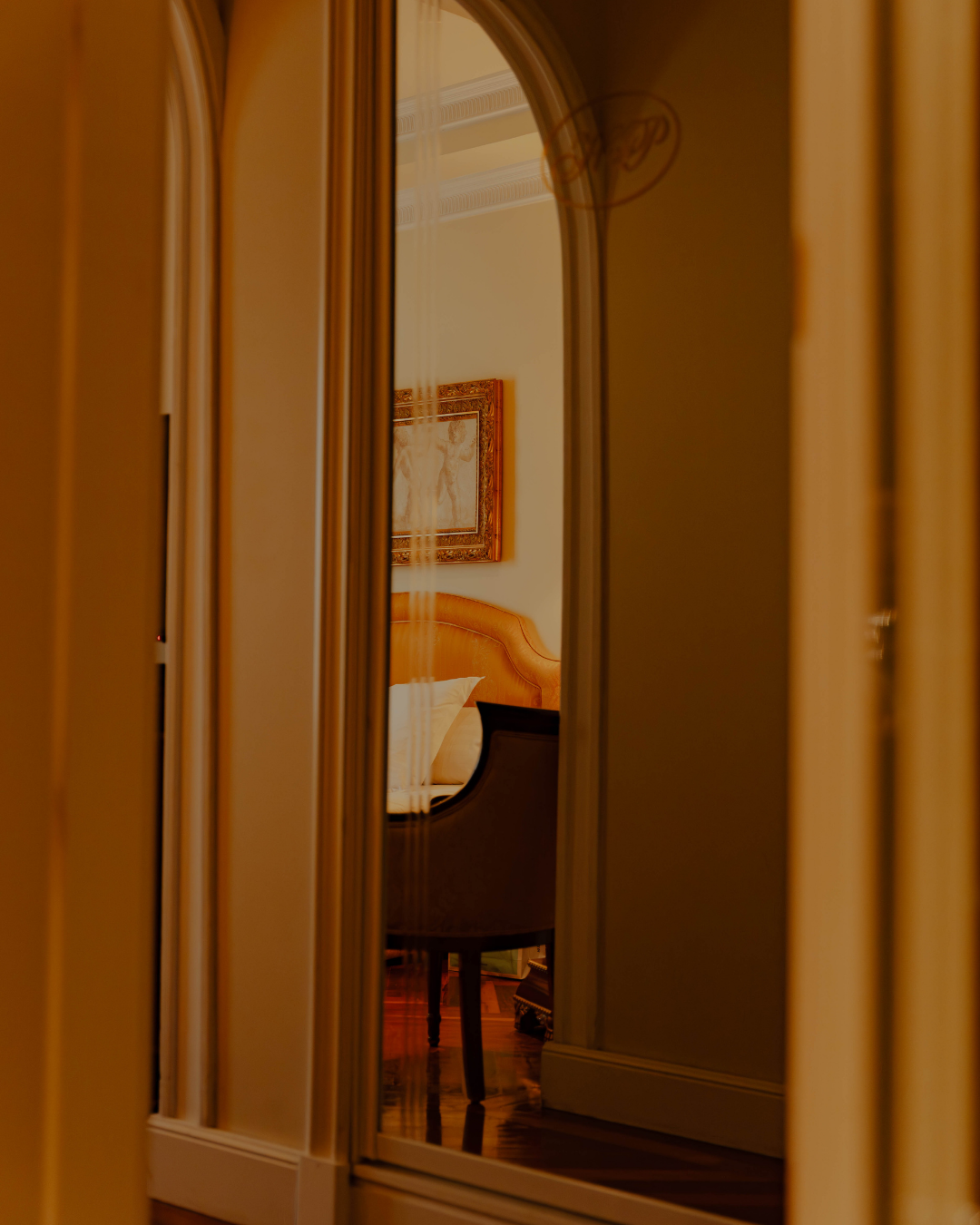 Une chambre vue à travers une porte miroir, avec un lit avec tête de lit en bois clair, un tableau au mur, et un fauteuil près du lit.