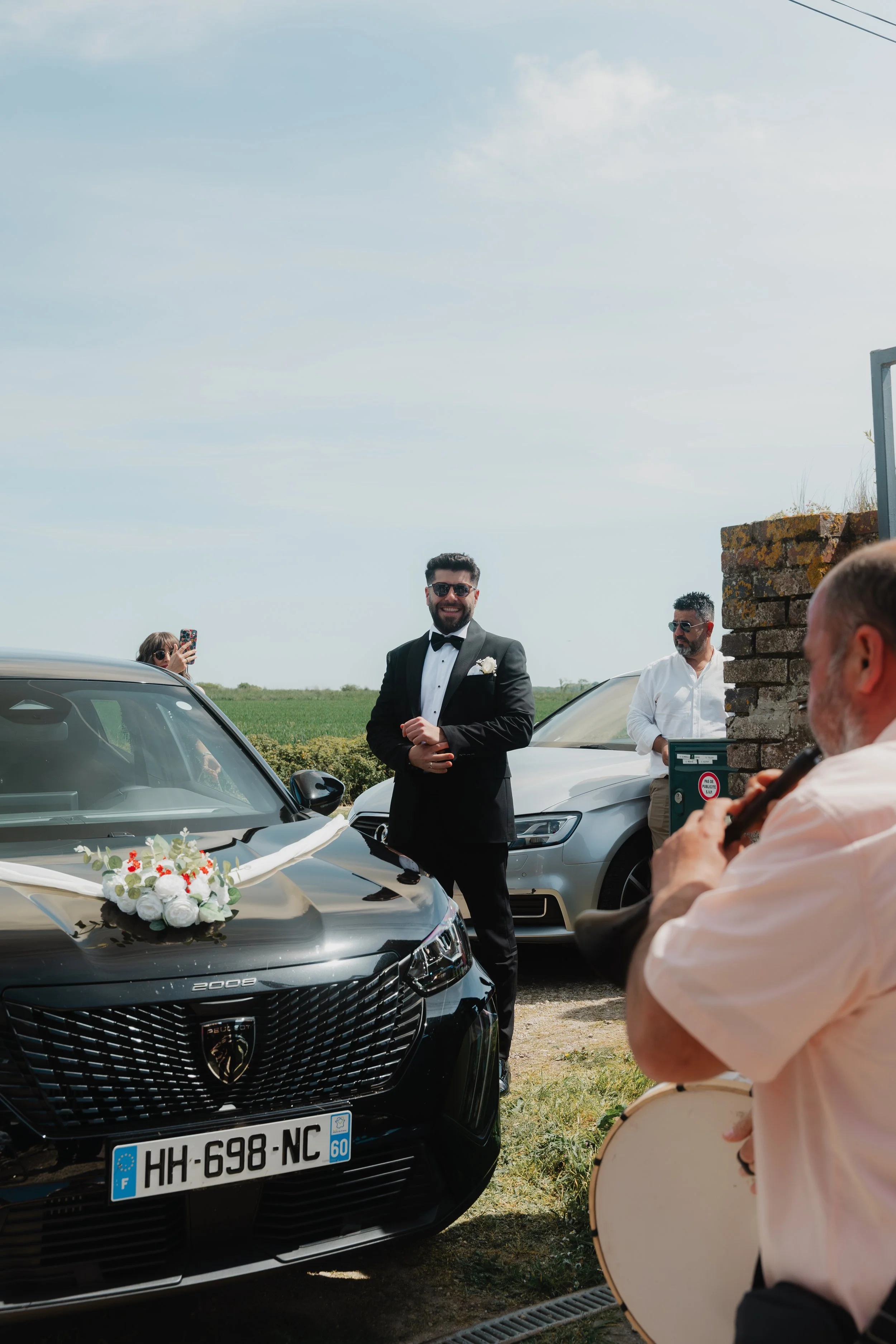 Un groupe de personnes lors d'une cérémonie de mariage en plein air, avec un homme en costume de mariage, une voiture décorée de fleurs en premier plan, et d'autres personnes en arrière-plan, dont une femme prenant une photo.