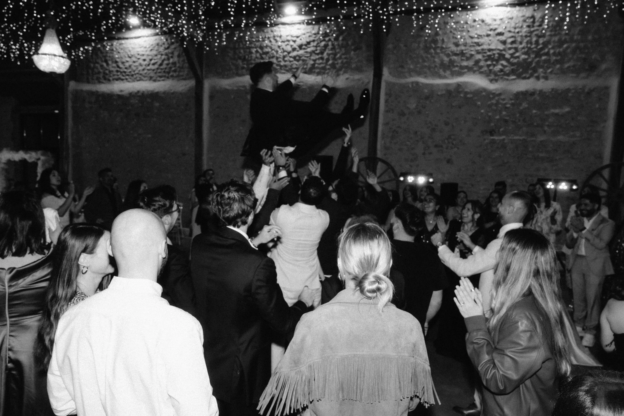 Une foule de personnes qui dansent lors d'une fête ou d'une célébration, avec une personne en costume suspendue en l'air au centre, sous un plafond décoré de guirlandes lumineuses.