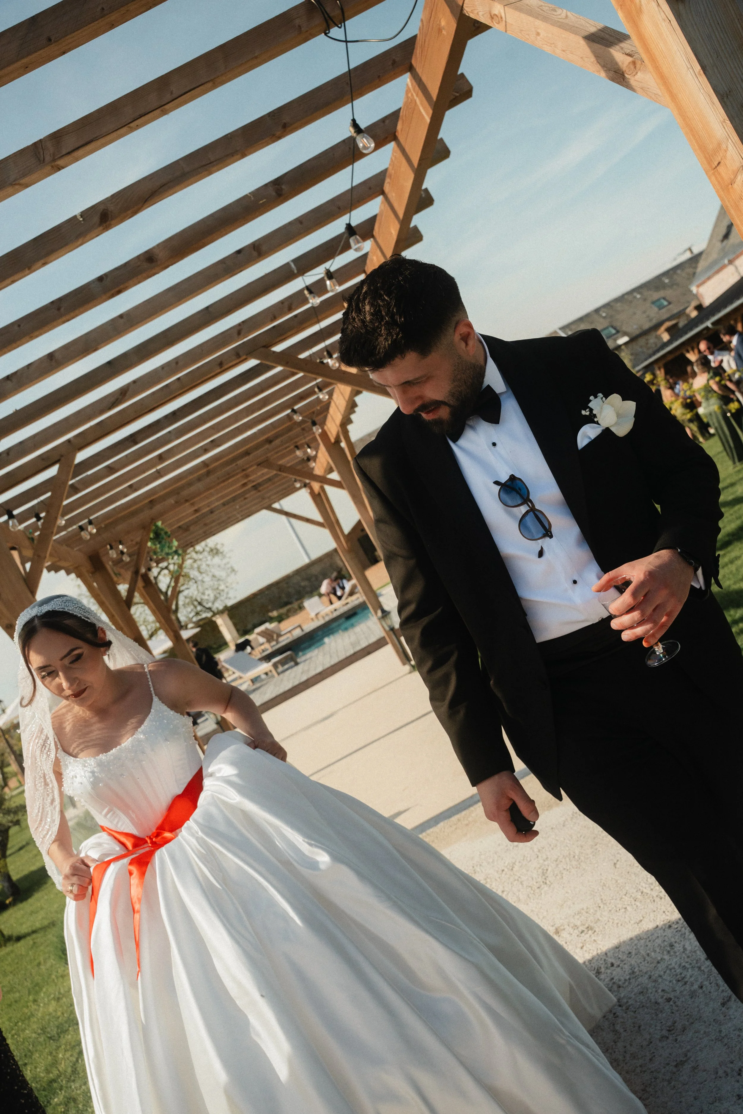 Un homme en smoking noir et une femme en robe de mariée blanche avec une ceinture orange lors d'un mariage en plein air.