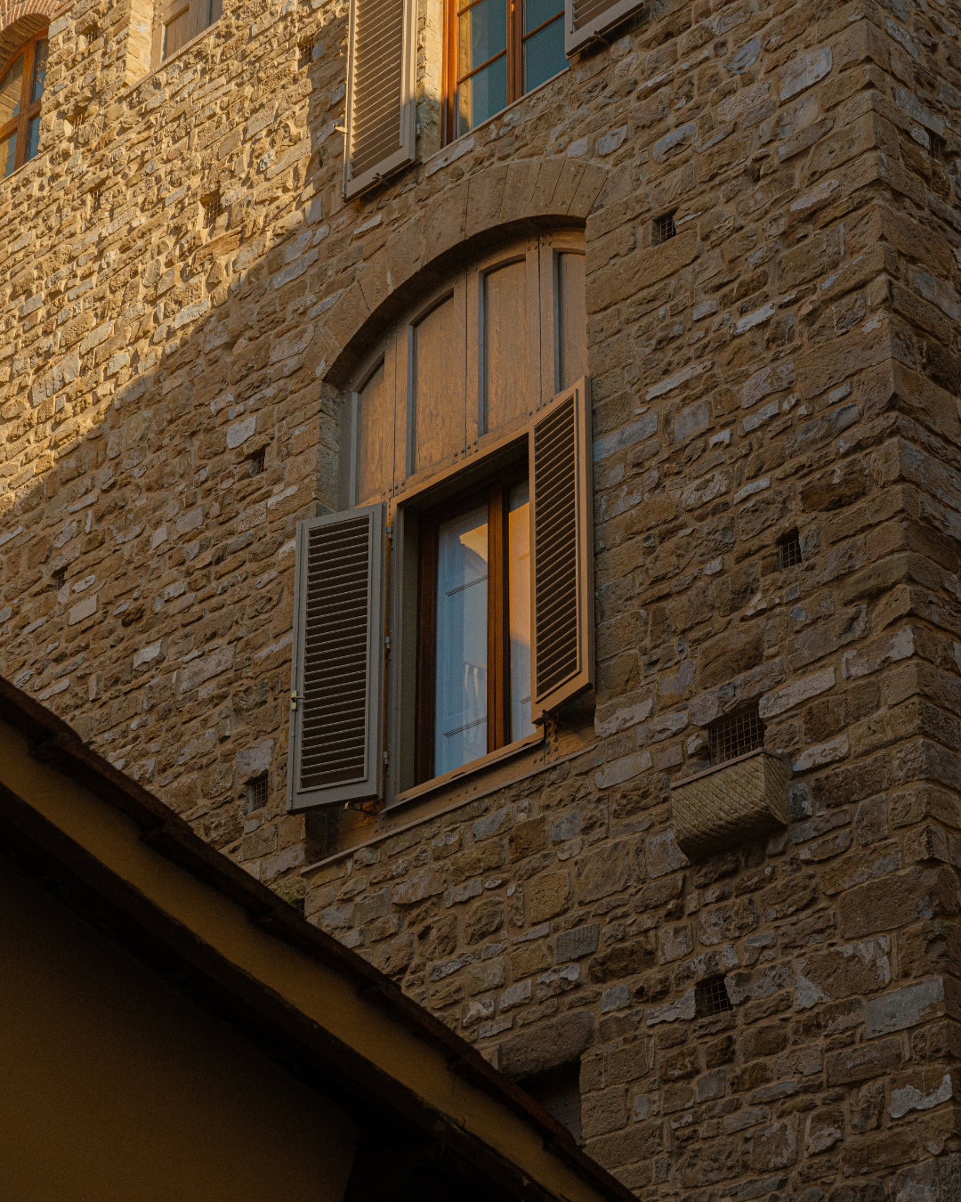 Fenêtre d'une maison en pierre avec volets en bois ouverts, reflet du coucher de soleil, murs en pierre beige